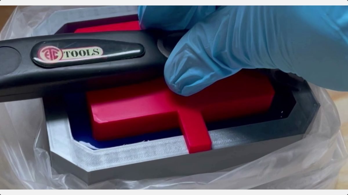 Blue-gloved hand holds a black clamp over the red printed soap master inside a gray mold base.