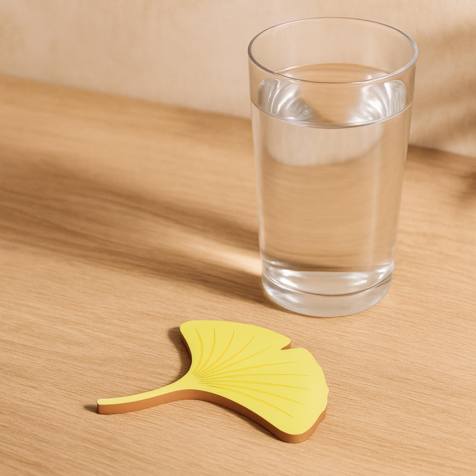 Yellow ginkgo leaf-shaped coaster on a wooden table beside a glass of water.