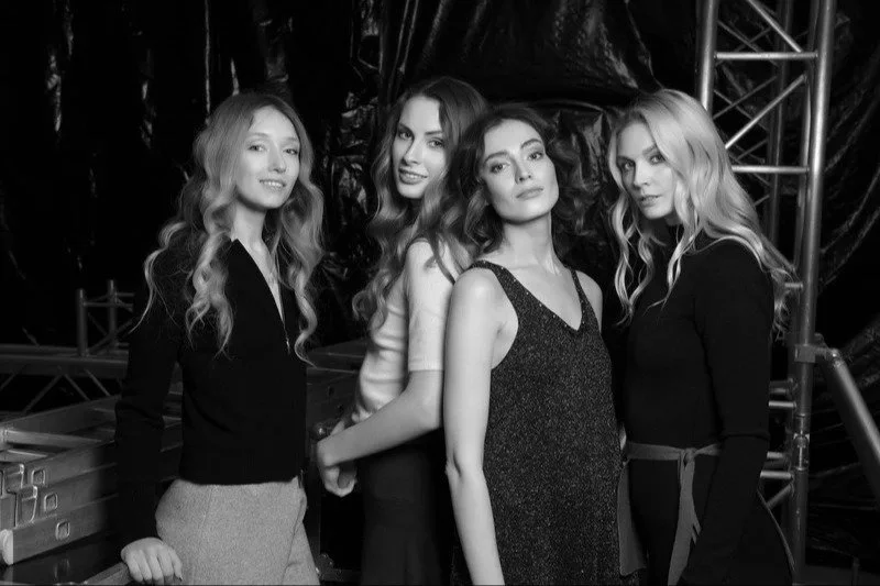 Black and white photo of four women standing together, posing and smiling, with a backdrop of industrial metal structures and a draped cloth.