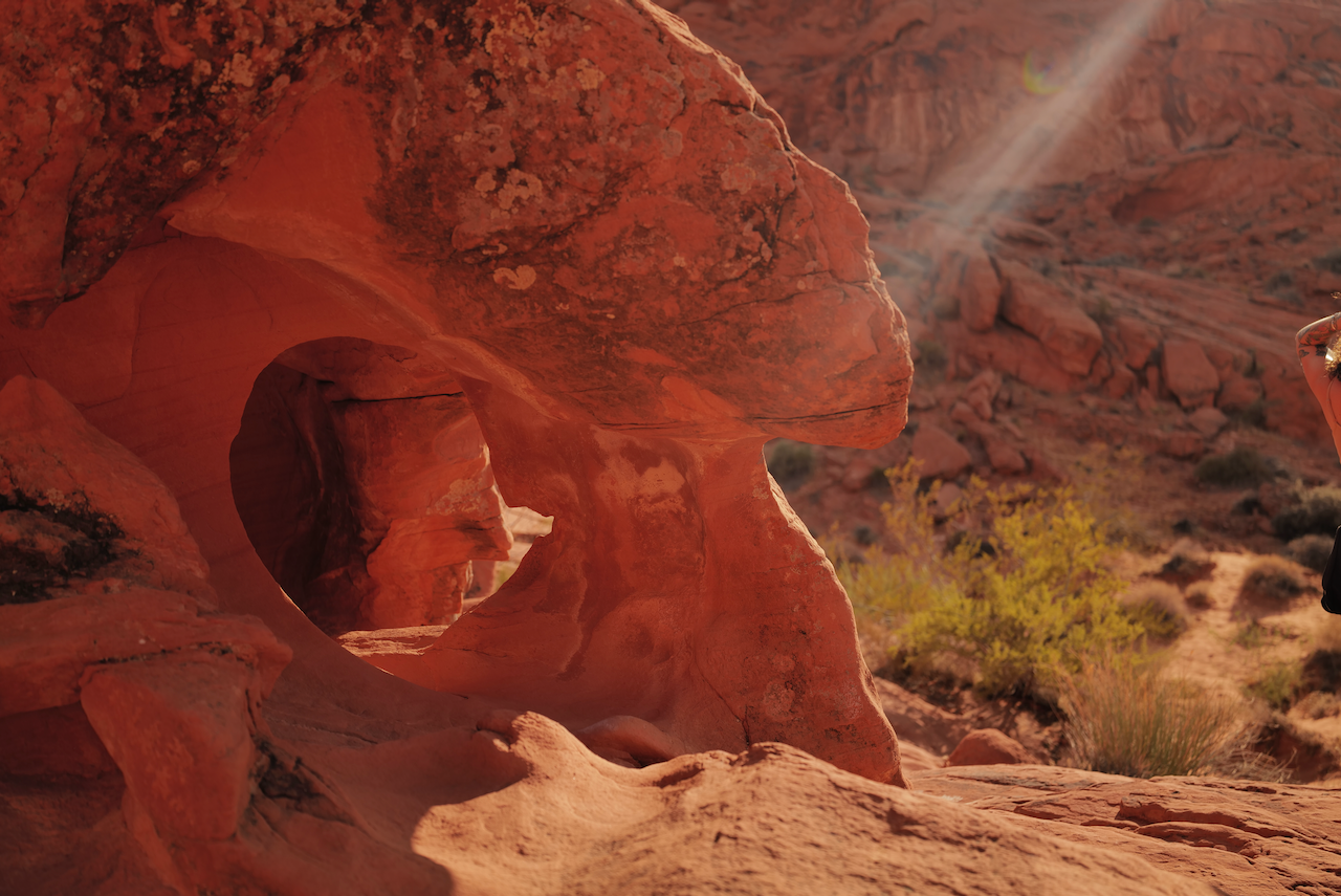 A desert landscape with red rock formations, including a round hole in the rock, with a small bush and shrubbery in the background.