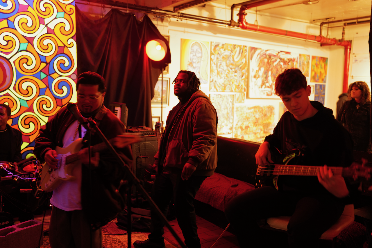 A band performs in an art gallery with colorful paintings and patterned artwork on the walls. The scene is lit with warm, red and orange lighting, creating an intimate atmosphere.