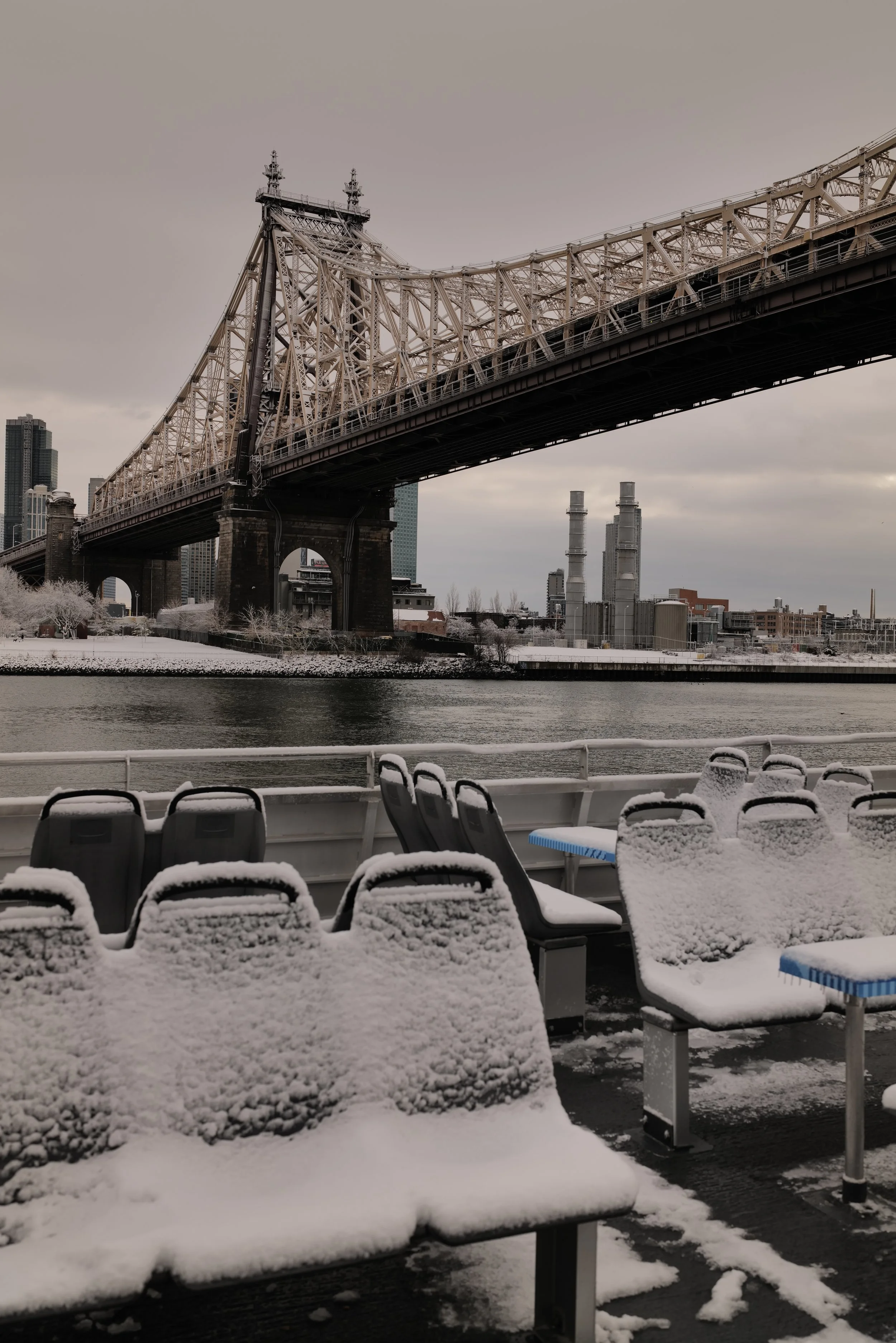 Snow-covered benches on a boat with the Queensboro Bridge and cityscape in the background.