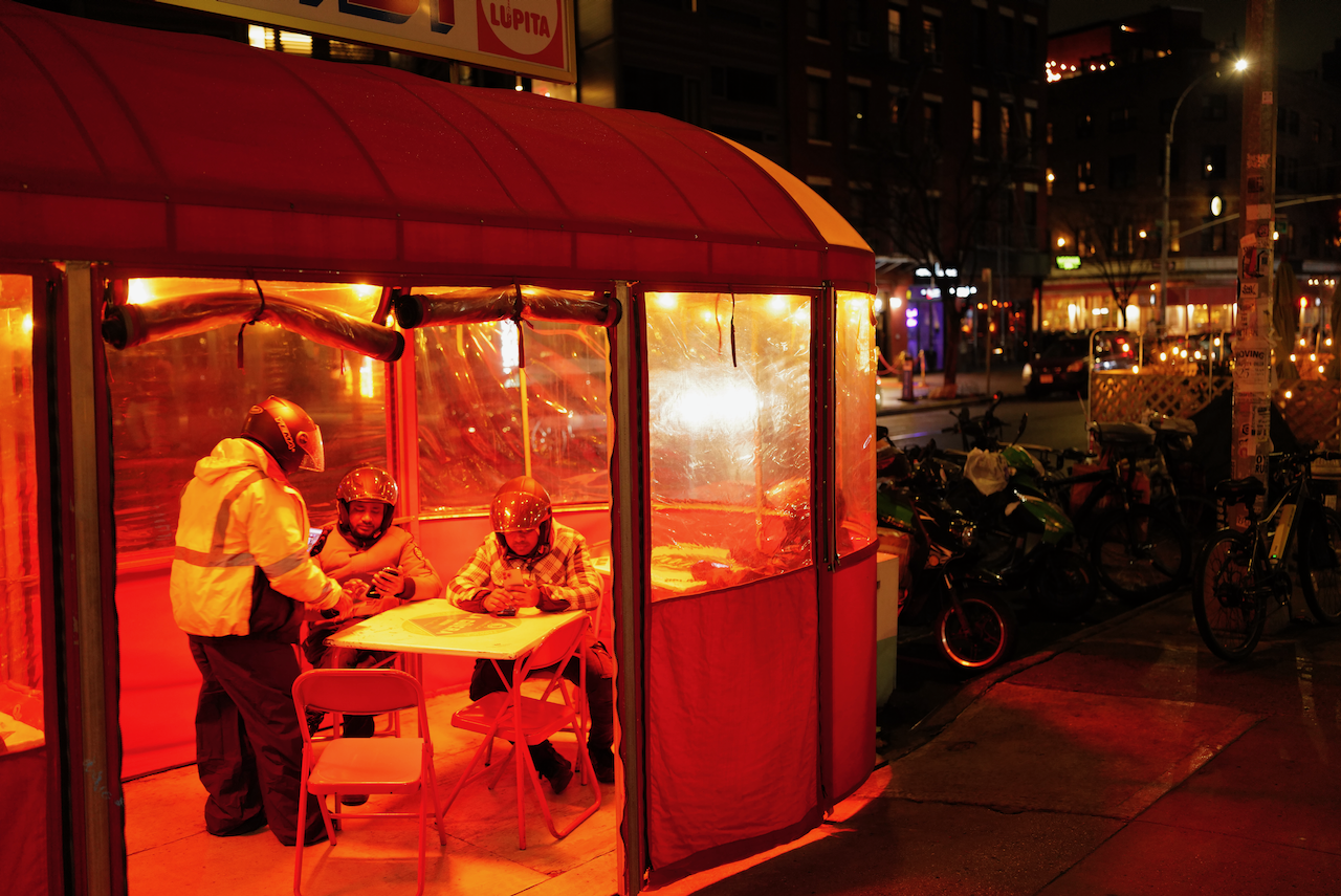 People sitting and standing inside a well-lit red tent on a city street at night, with bicycles parked outside. The scene is illuminated by streetlights and nearby buildings.