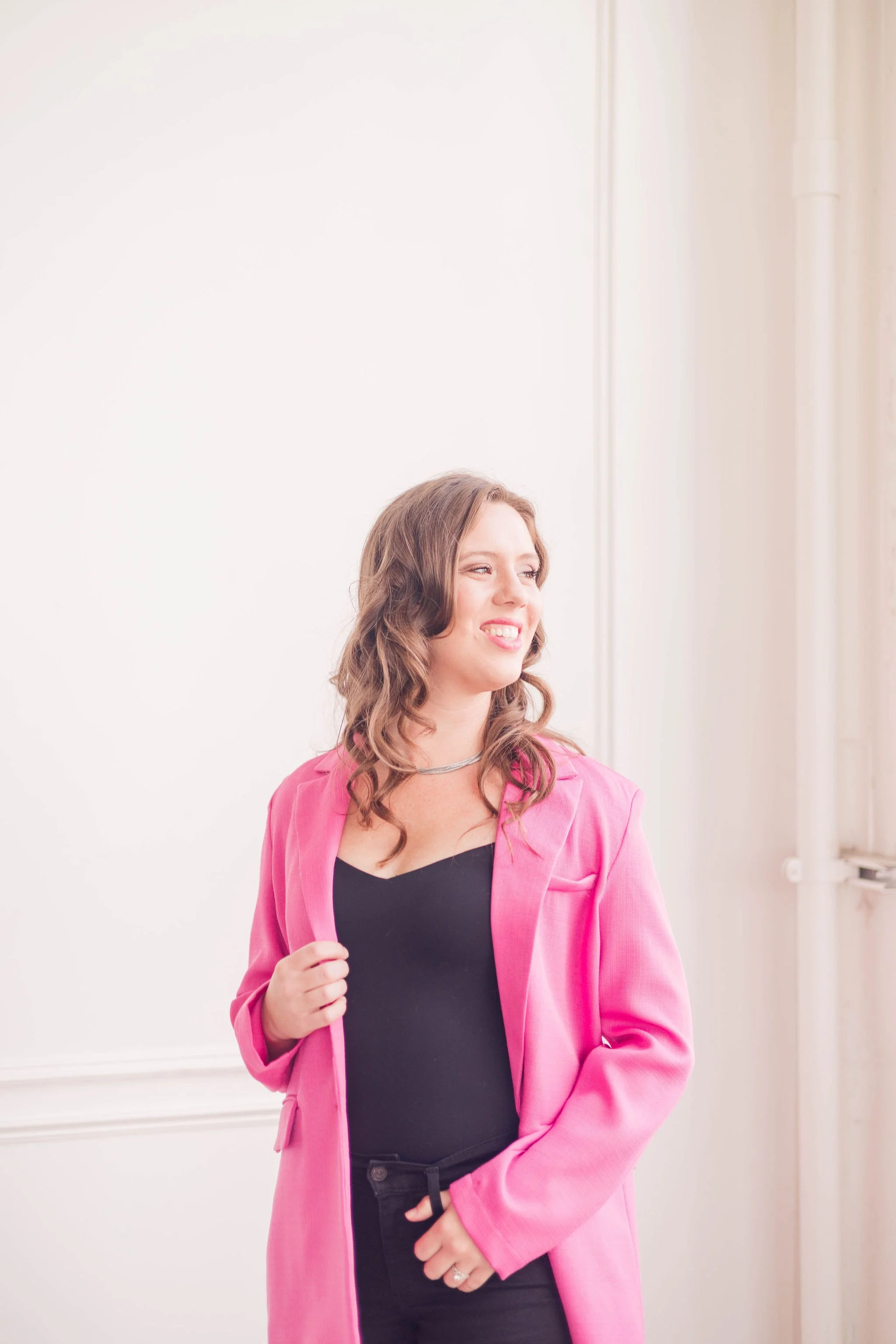 A young woman with wavy brown hair smiling while wearing a pink blazer over a black top, standing indoors near a white wall.