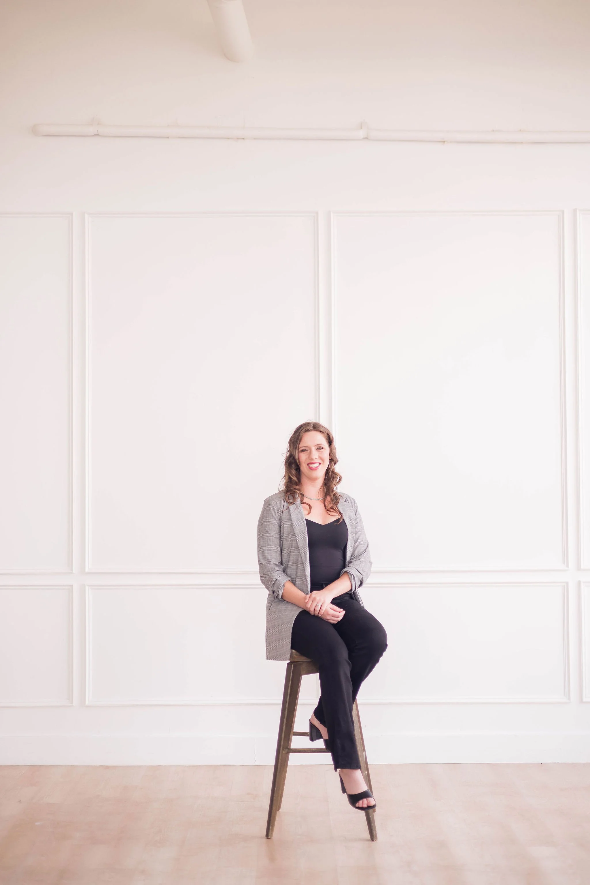 A woman sitting on a stool in a minimal white room with decorative wall panels, wearing a gray blazer, black top, black pants, and black high heels, smiling.