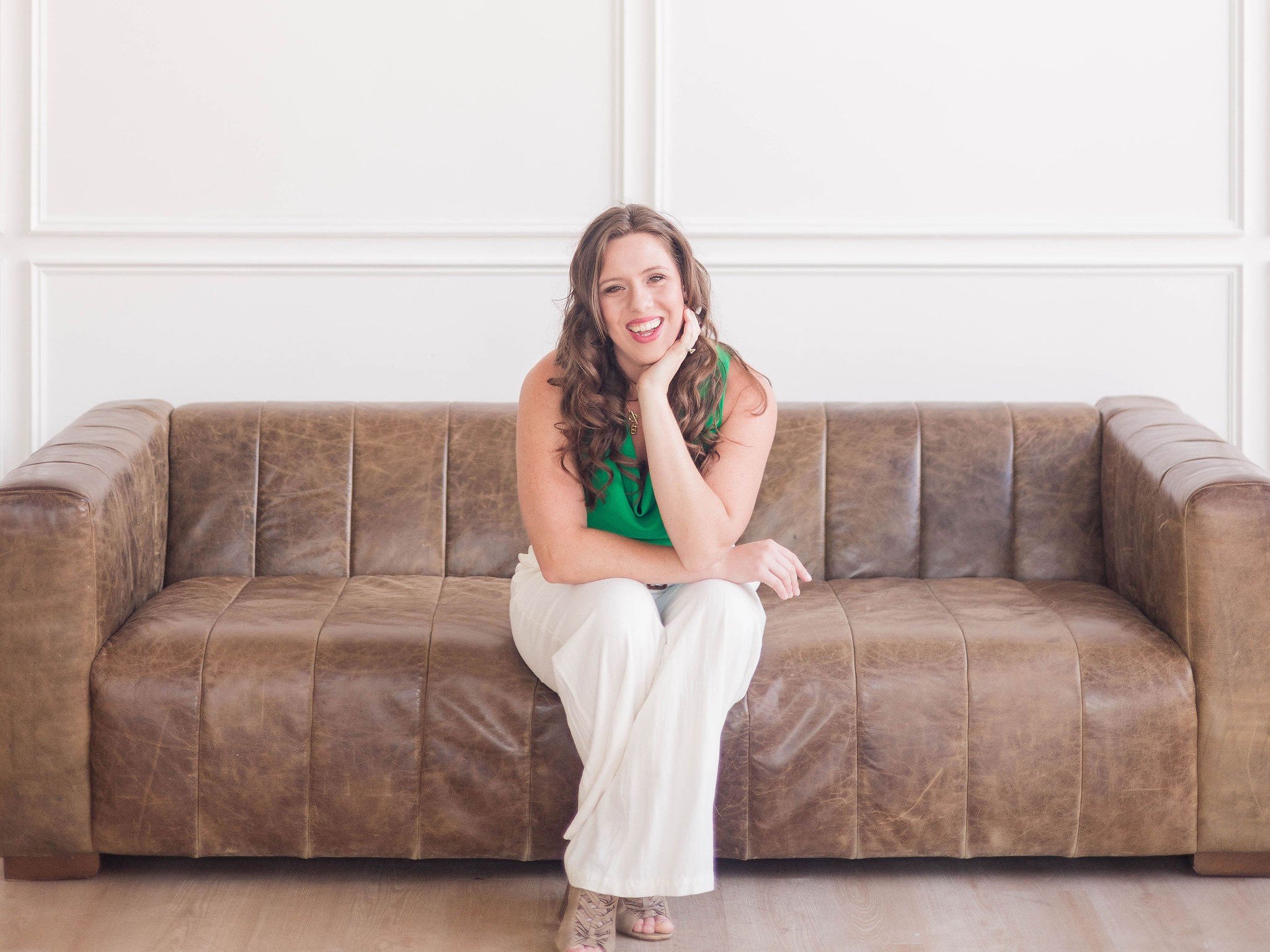 A young woman with long curly brown hair sitting on a brown leather sofa, wearing a green sleeveless top and white pants, smiling with her head resting on her hand in a bright room with white paneled walls.