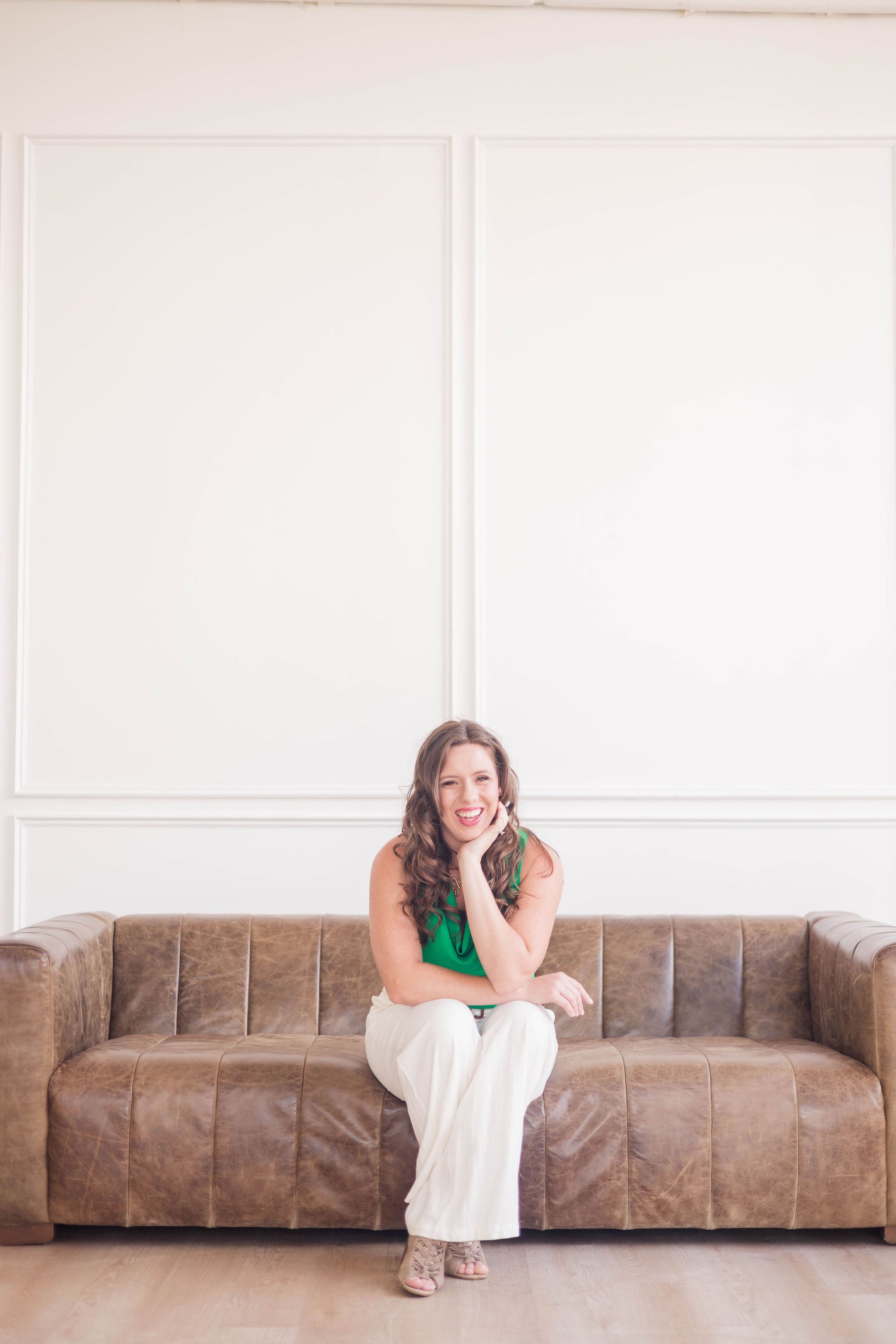 A woman with brown curly hair sitting on a brown leather sofa, smiling, wearing a green top and white pants, in a bright room with white walls.