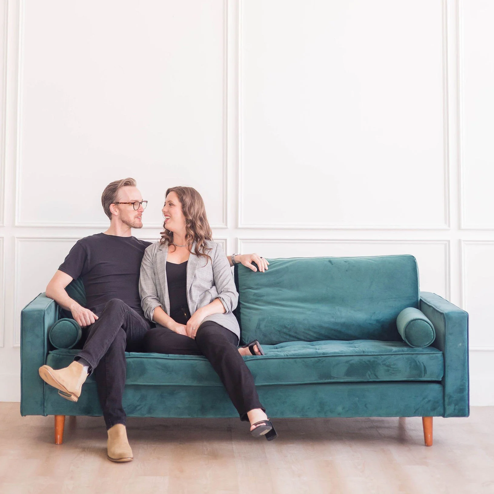 A man and woman sitting close together on a green velvet couch, smiling and looking into each other's eyes in a bright room with white paneled walls.