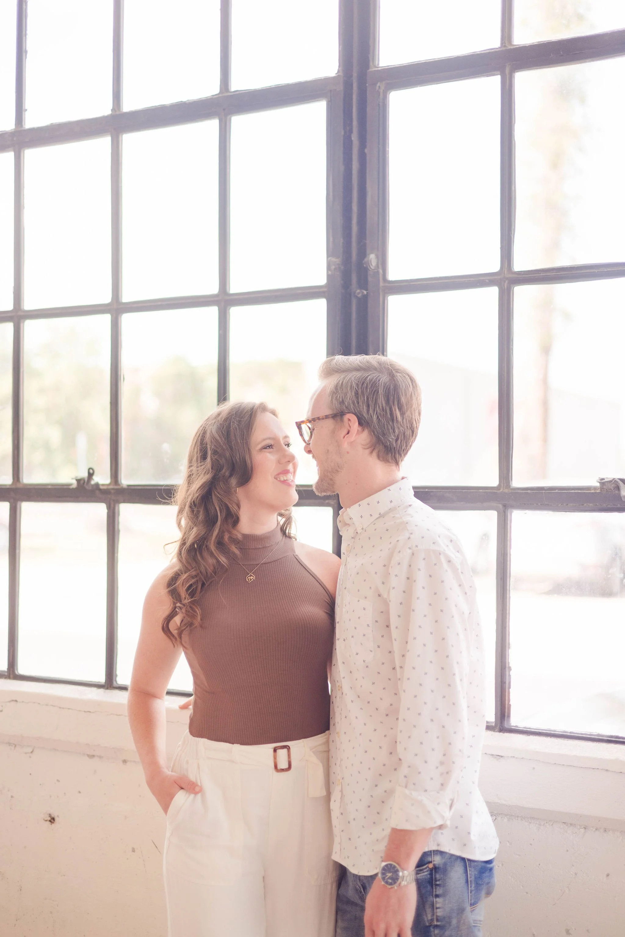 A couple standing close together inside, smiling and looking into each other's eyes near a large window.