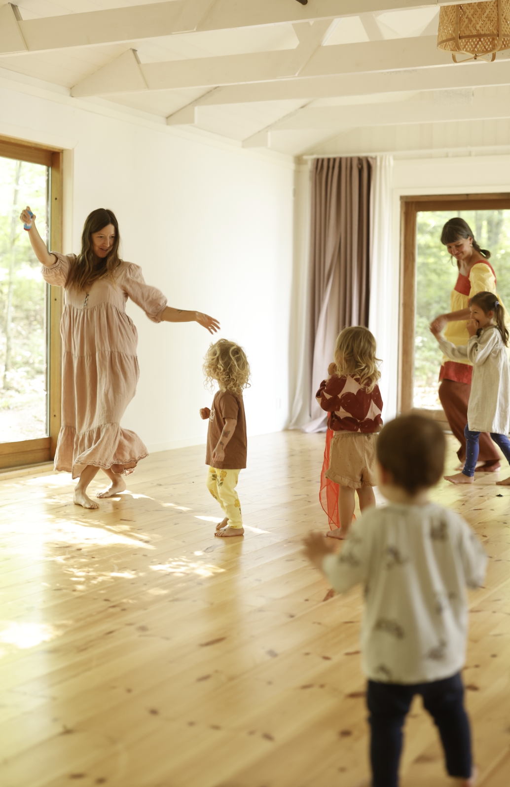 A woman dancing and playing with children indoors, with others in the background, sunlight streaming through large windows.