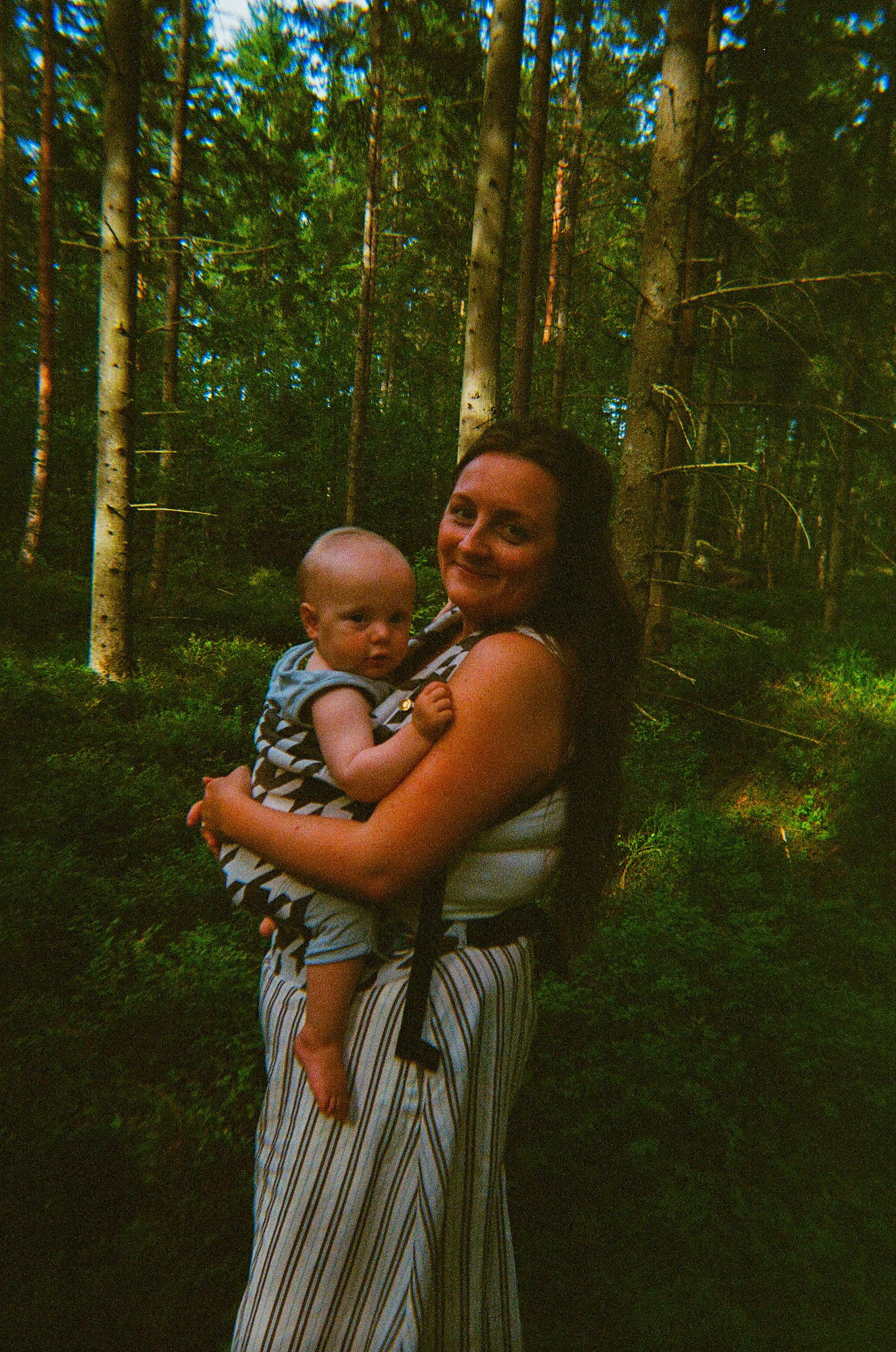 A woman holding a baby in a forest with tall trees and green foliage.