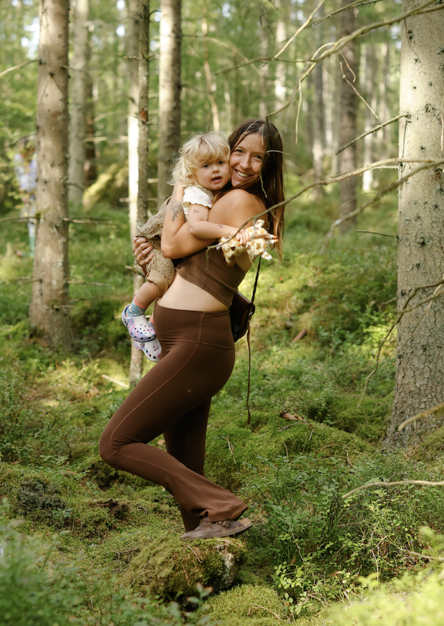 A woman carrying a small child on her back in a forest with green trees and moss-covered ground, smiling and looking at the camera.