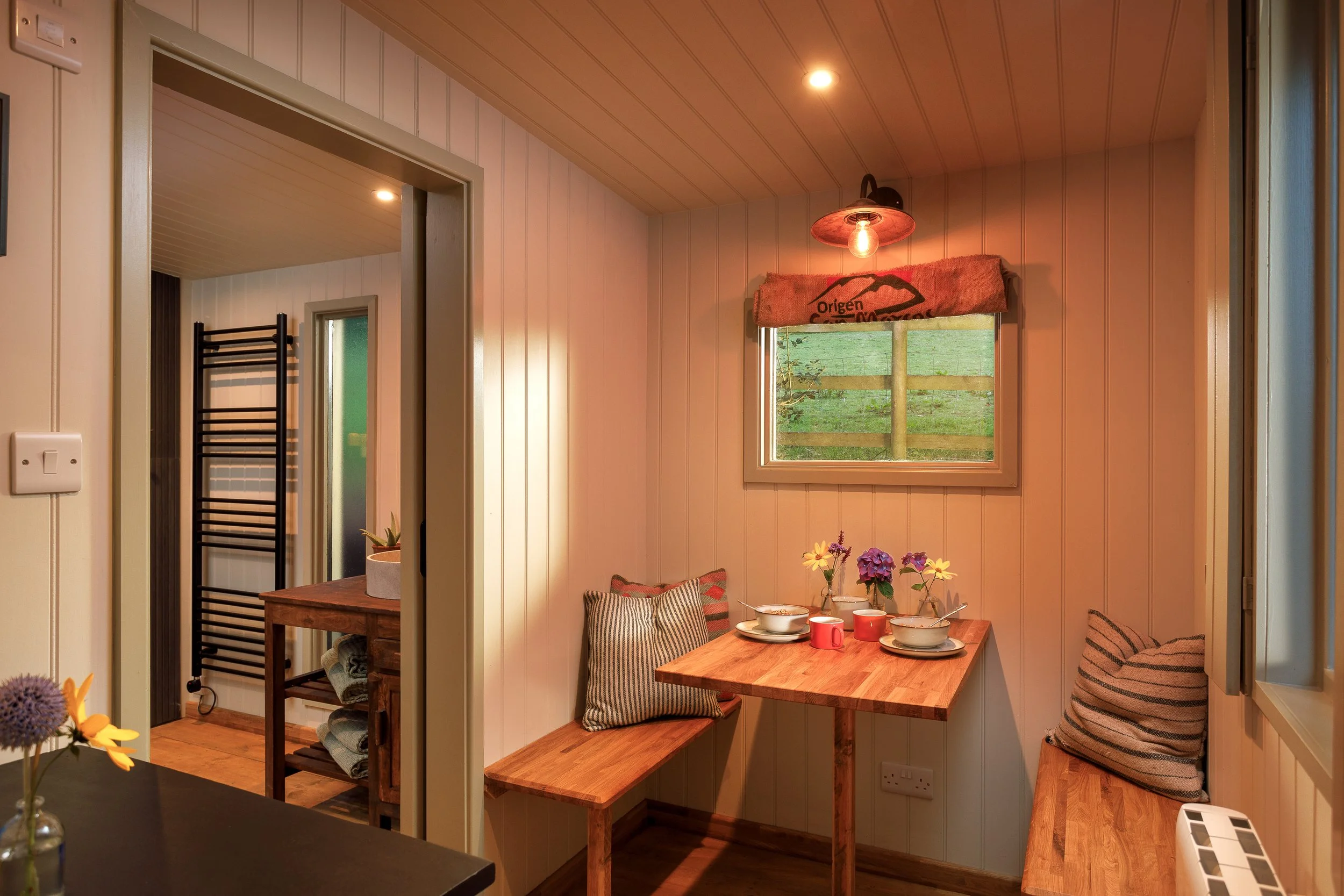 A cozy dining nook with a wooden table set for breakfast, surrounded by striped pillows and small vases of flowers, with a window showing greenery outside and warm ambient lighting.