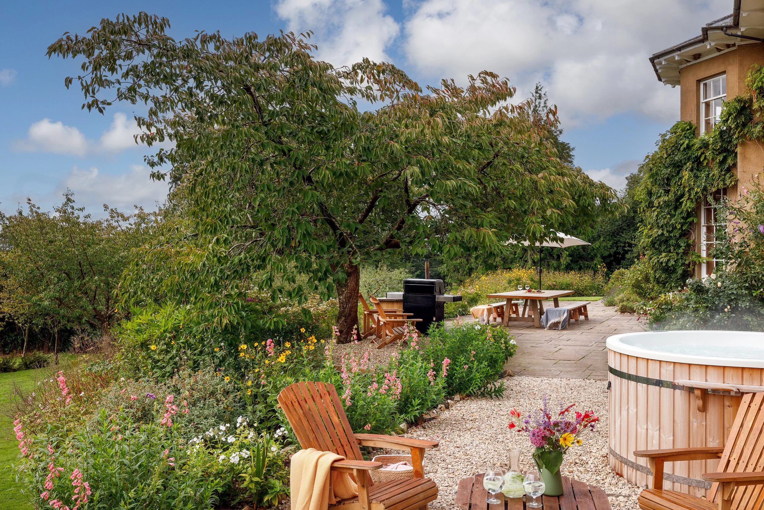 A backyard patio with garden furniture, a hot tub, a table with a flower vase, a barbecue grill, and surrounding lush greenery and flowers under a partly cloudy sky.