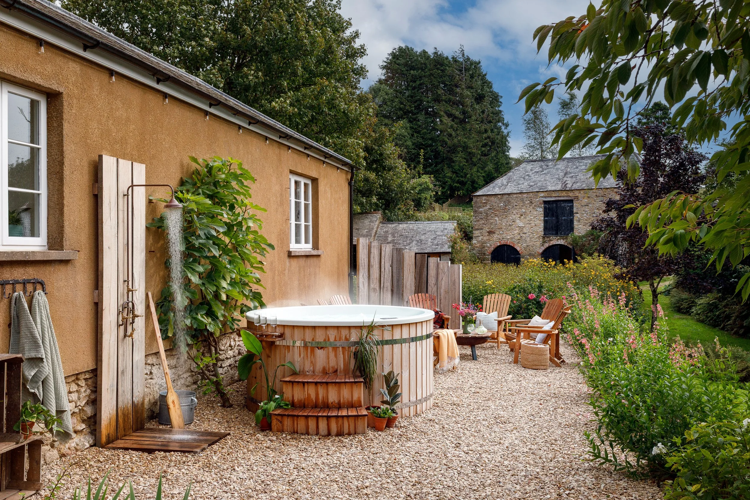 Outdoor garden with a hot tub, wooden chairs, colorful flowers, and a rustic barn in the background.