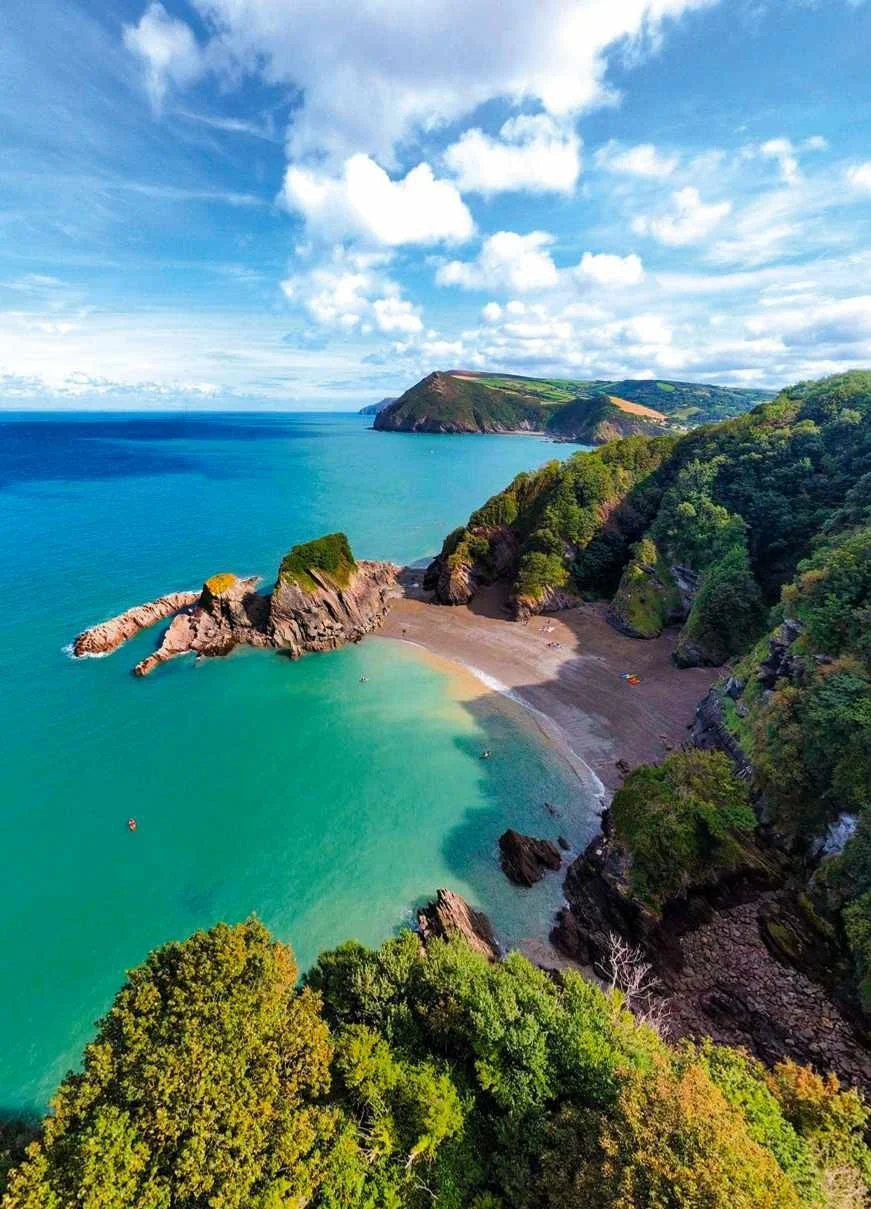 A scenic coastal landscape with blue ocean, sandy beach, rocky formations, and lush green hills under a partly cloudy sky.