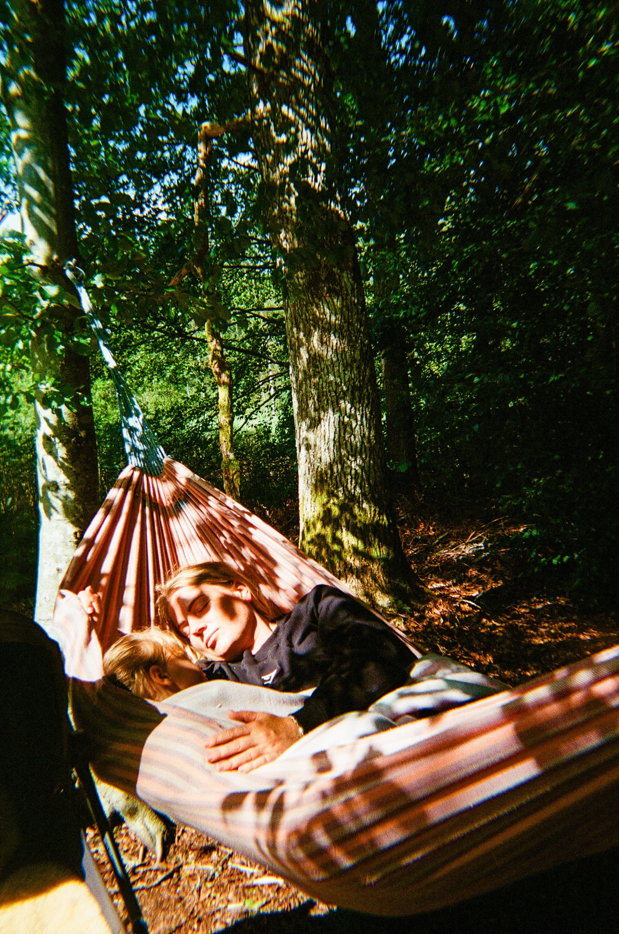 A young man and woman lying in a hammock outdoors among trees, sharing a tender moment.