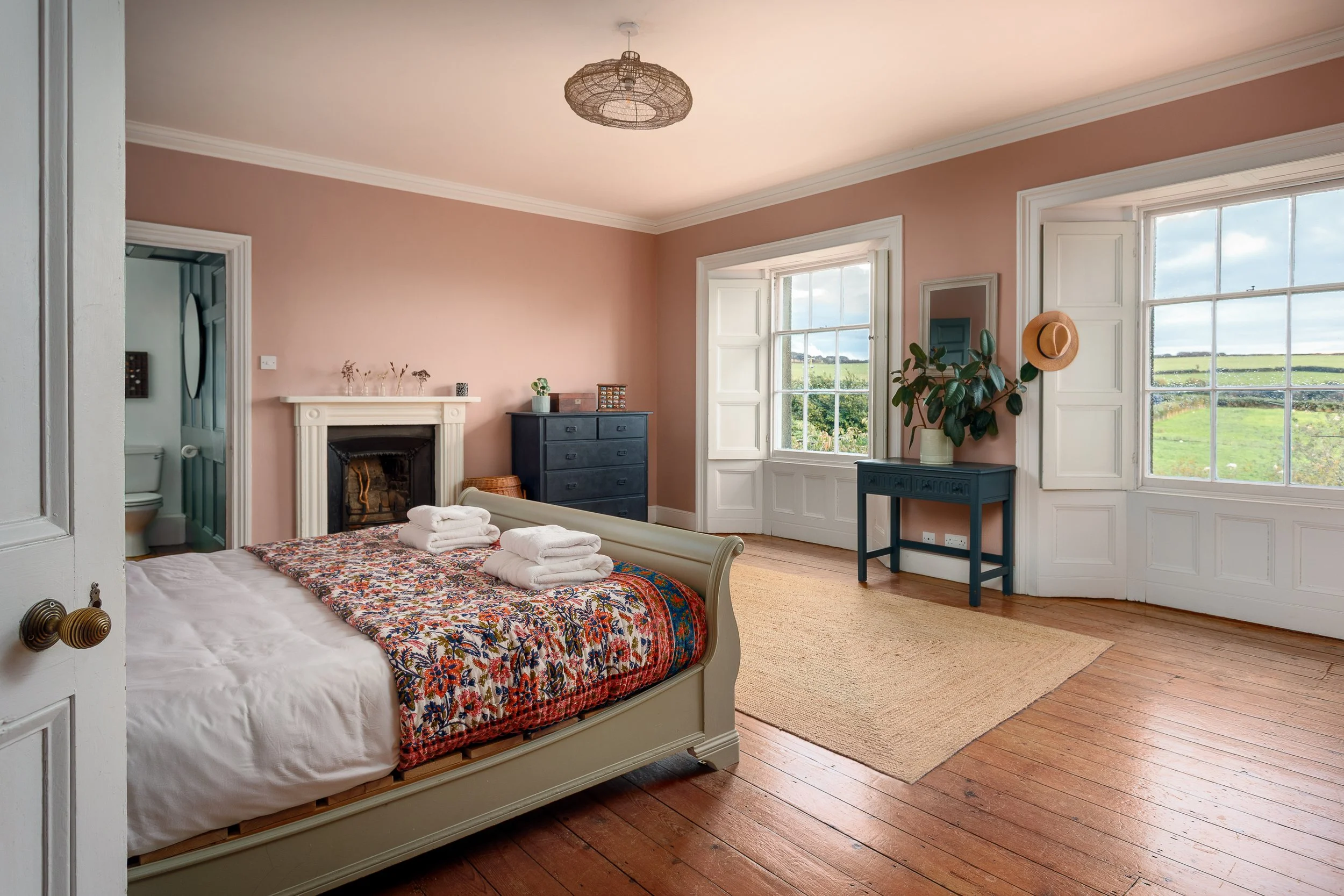 A bedroom with wooden floors, light pink walls, a white bed with a colorful quilt, and folded towels on the bed. There are large windows with white shutters, a dark blue dresser, a small blue table with a plant and a hat, and an entrance to a bathroom with a toilet. A woven ceiling light hangs from the ceiling.