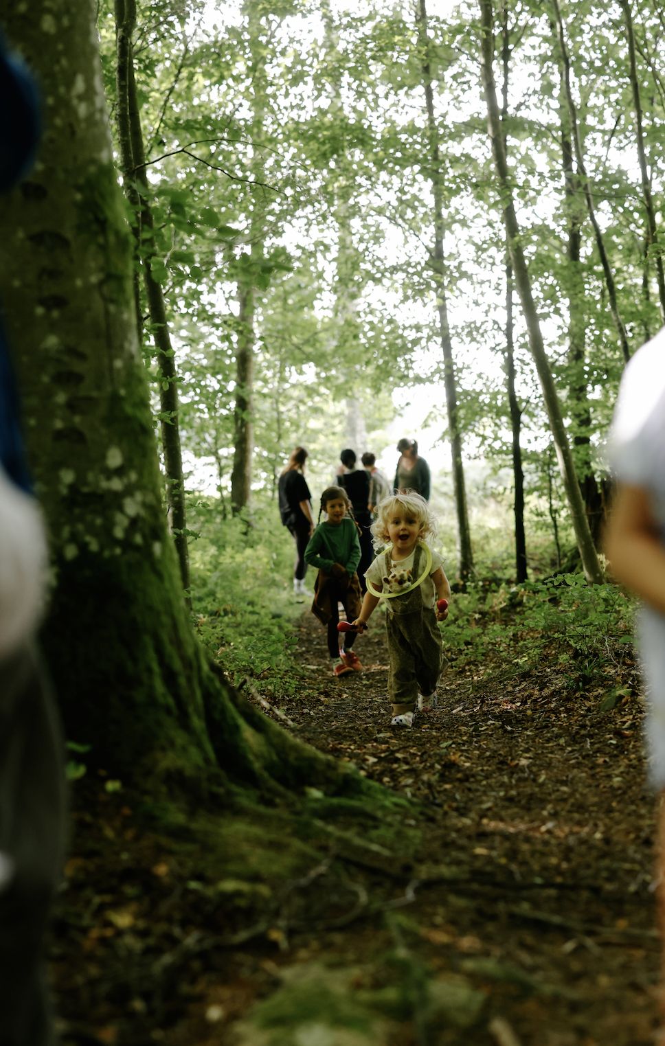 Children playing outdoors on a forest trail, with a young girl smiling and holding a toy, surrounded by trees and additional children and adults in the background.