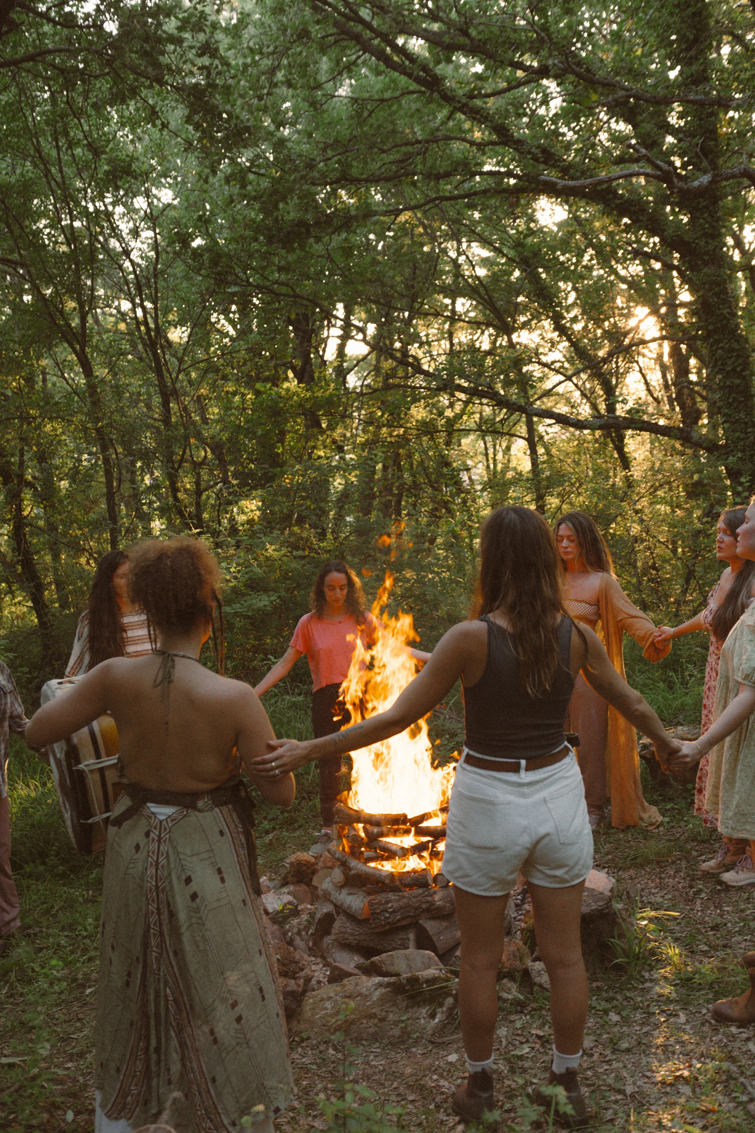 A group of people holding hands in a circle around a campfire in a wooded area during sunset.