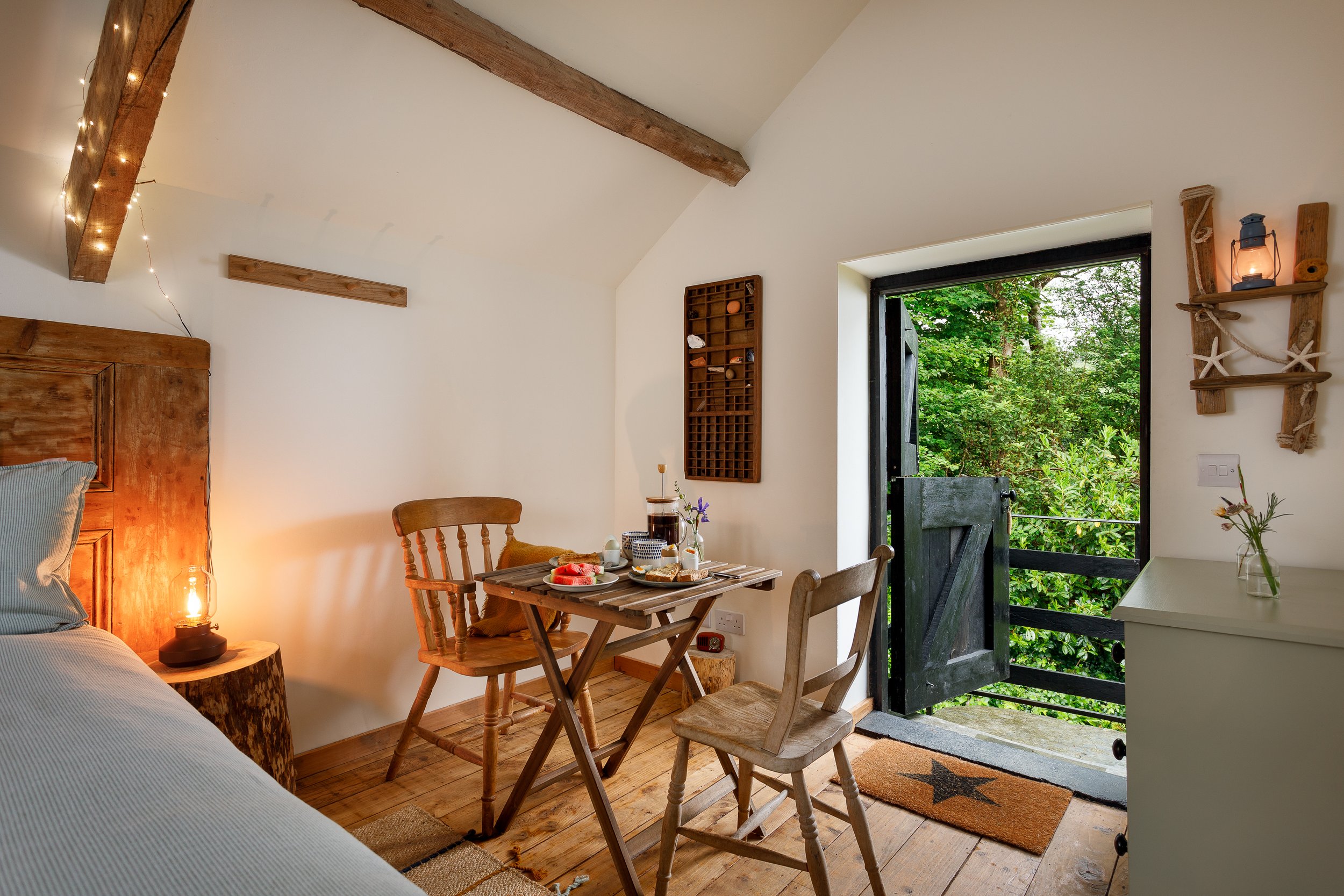 Cozy bedroom with wooden furniture, small table set for breakfast, open door to a green outdoor view, decorated with rustic shelves and string lights.