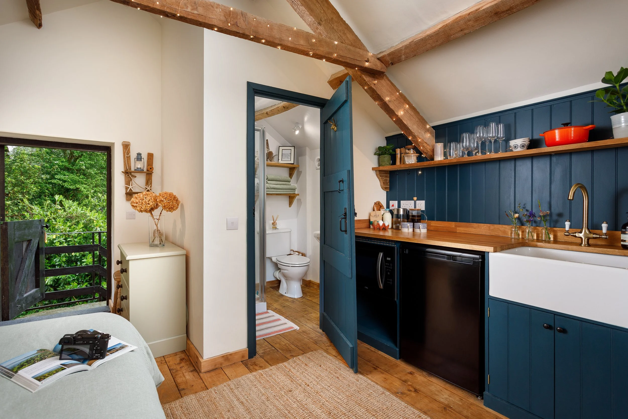 Interior of a cozy cottage or small home with a view of a kitchen and bathroom. The kitchen has blue cabinets, a wooden countertop, a white farmhouse sink, and open shelves with glassware and plants. Visible wooden beams in the ceiling are decorated 