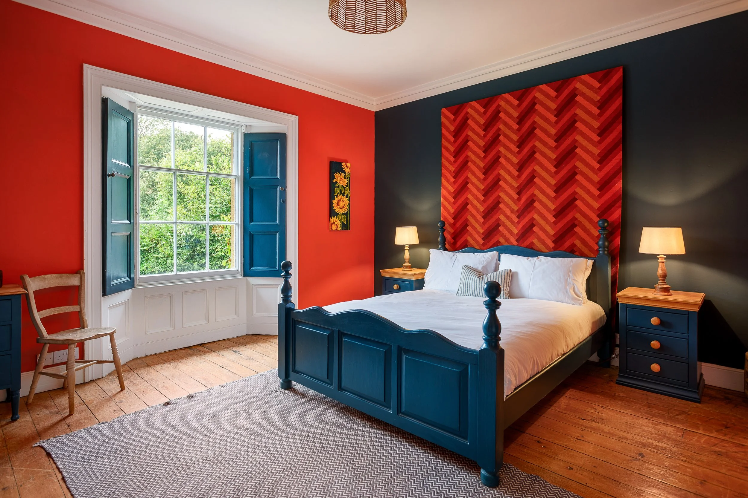 A bedroom with red and dark blue walls, a large window with blue shutters, a blue bed frame with white bedding, and two nightstands with lamps. There is a wooden chair near the window and a wooden floor, with a patterned rug.