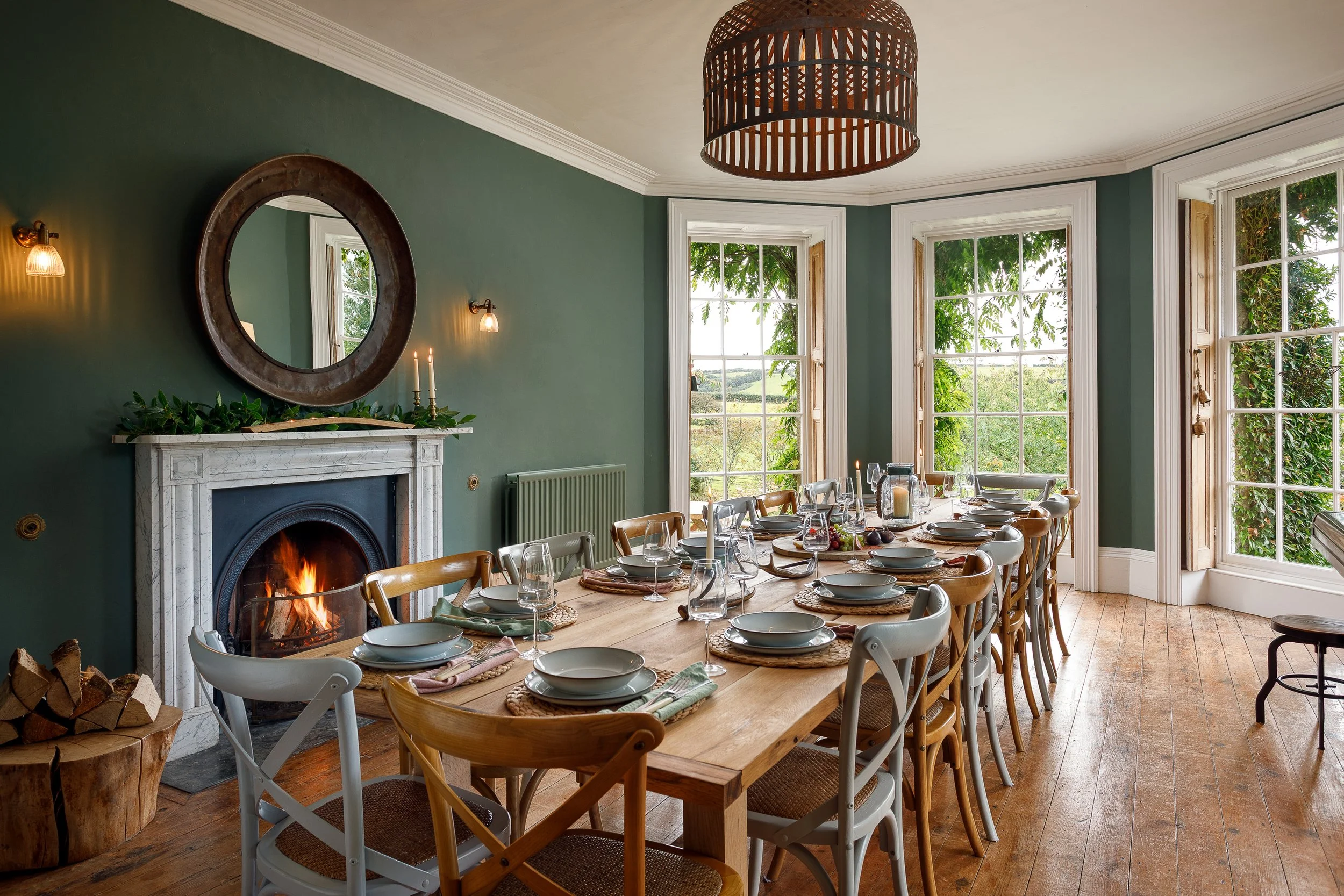 A dining room with a long wooden table set with plates, glasses, candles, and napkins, near large windows with green outdoor scenery, a fireplace with a fire, and a hanging wooden light fixture.