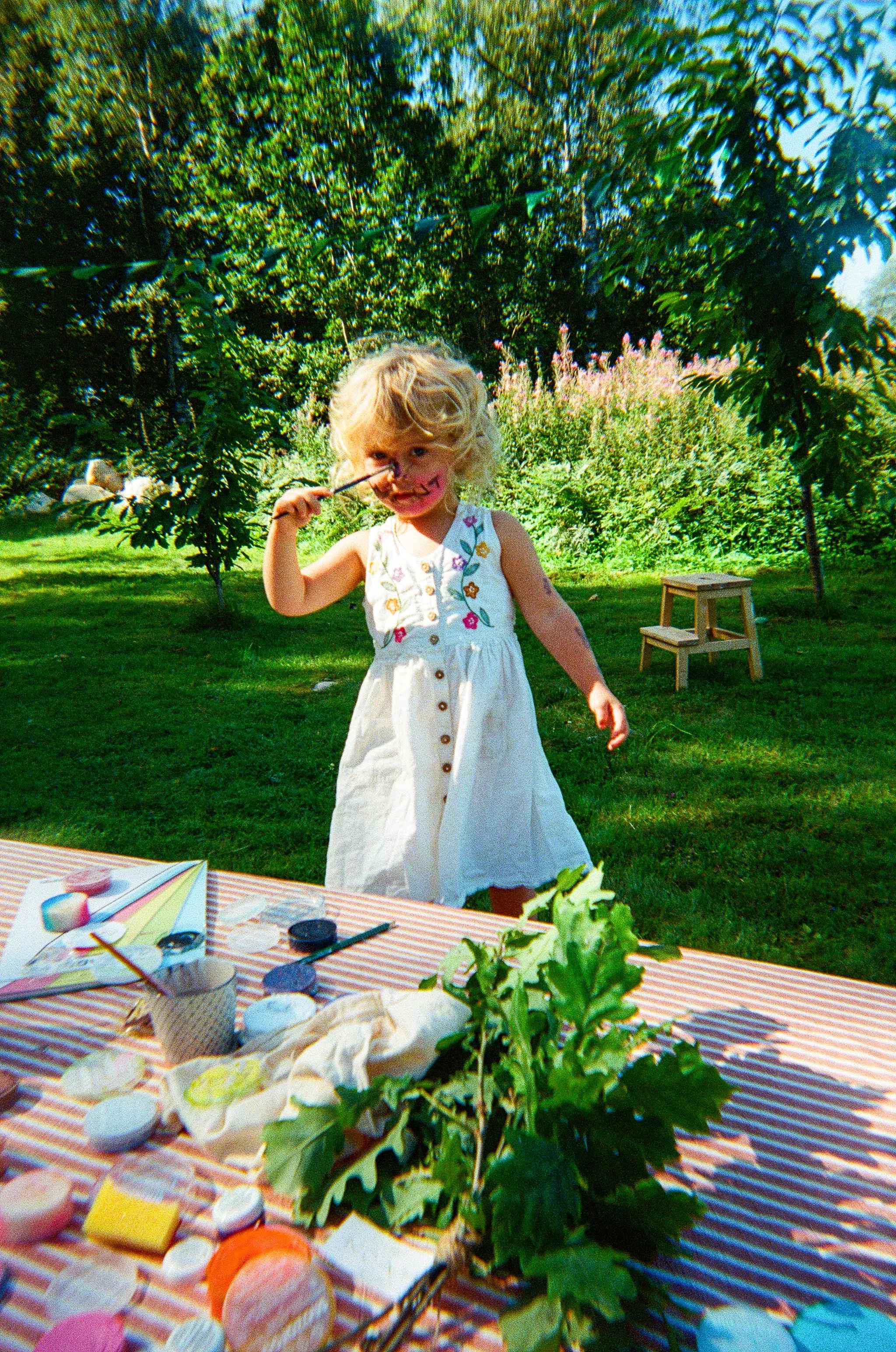 A young girl with blond curly hair, wearing a white dress with colorful embroidery, paints her face with a pink and black design, standing outdoors in a lush green garden. A table in the foreground has various face paint supplies and a bunch of green