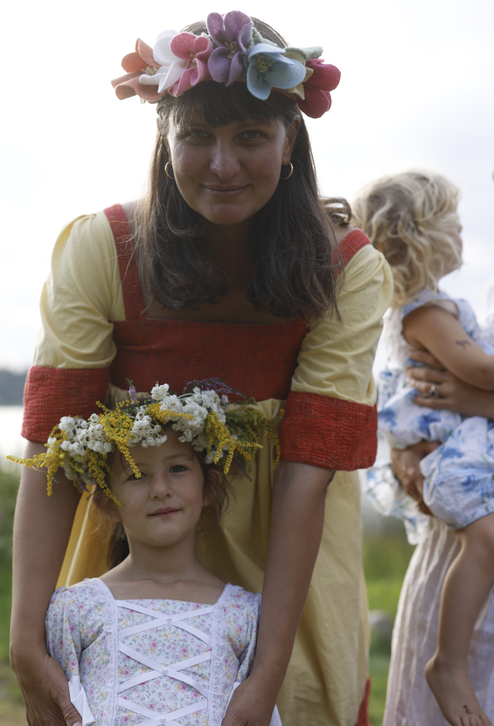 Woman in a yellow dress with red accents and a floral crown, smiling as she leans over a young girl wearing a floral dress and a flower crown, outdoors during daytime.