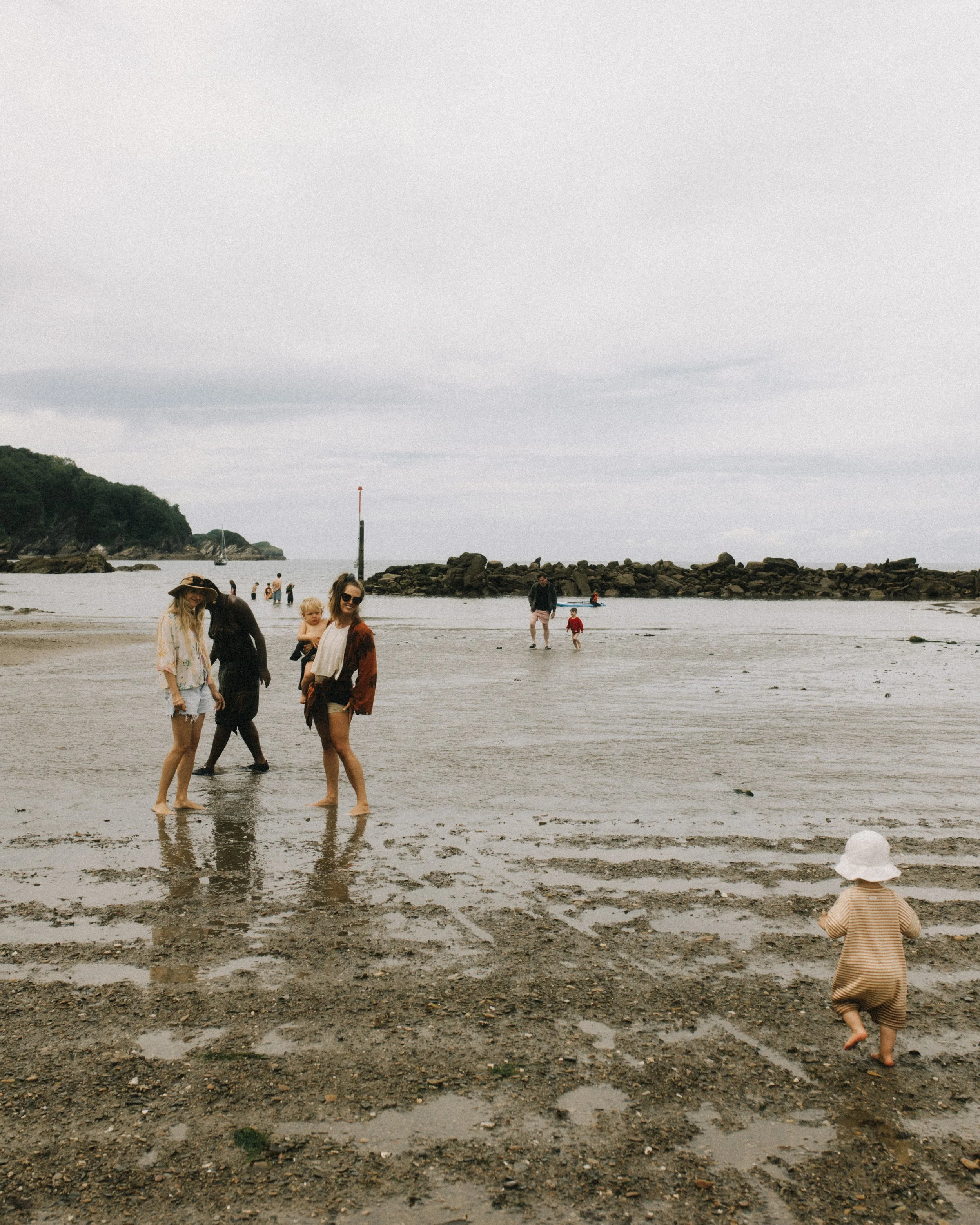 People enjoying a day at the beach, with children and adults walking on the wet sand near rocks, a boat with a sailboat in the background, and a cloudy sky overhead.