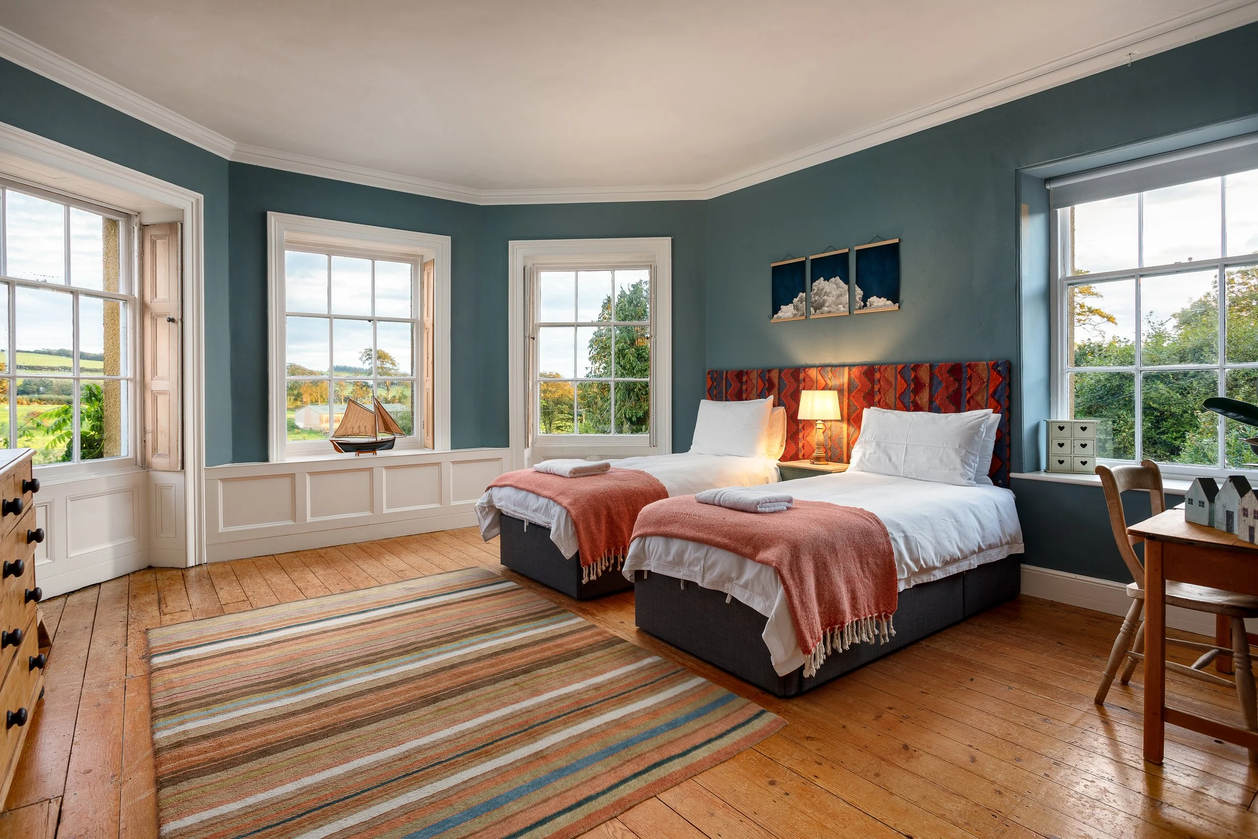 A spacious bedroom with three large windows, two twin beds with red patterned headboards, white bedding, pink throws, and folded towels. There's a wooden dresser, a small table with house-shaped decorations, and a framed cloud artwork above the beds. The room has wooden floors, a striped rug, a small lamp, and views of green trees and a cloudy sky outside.