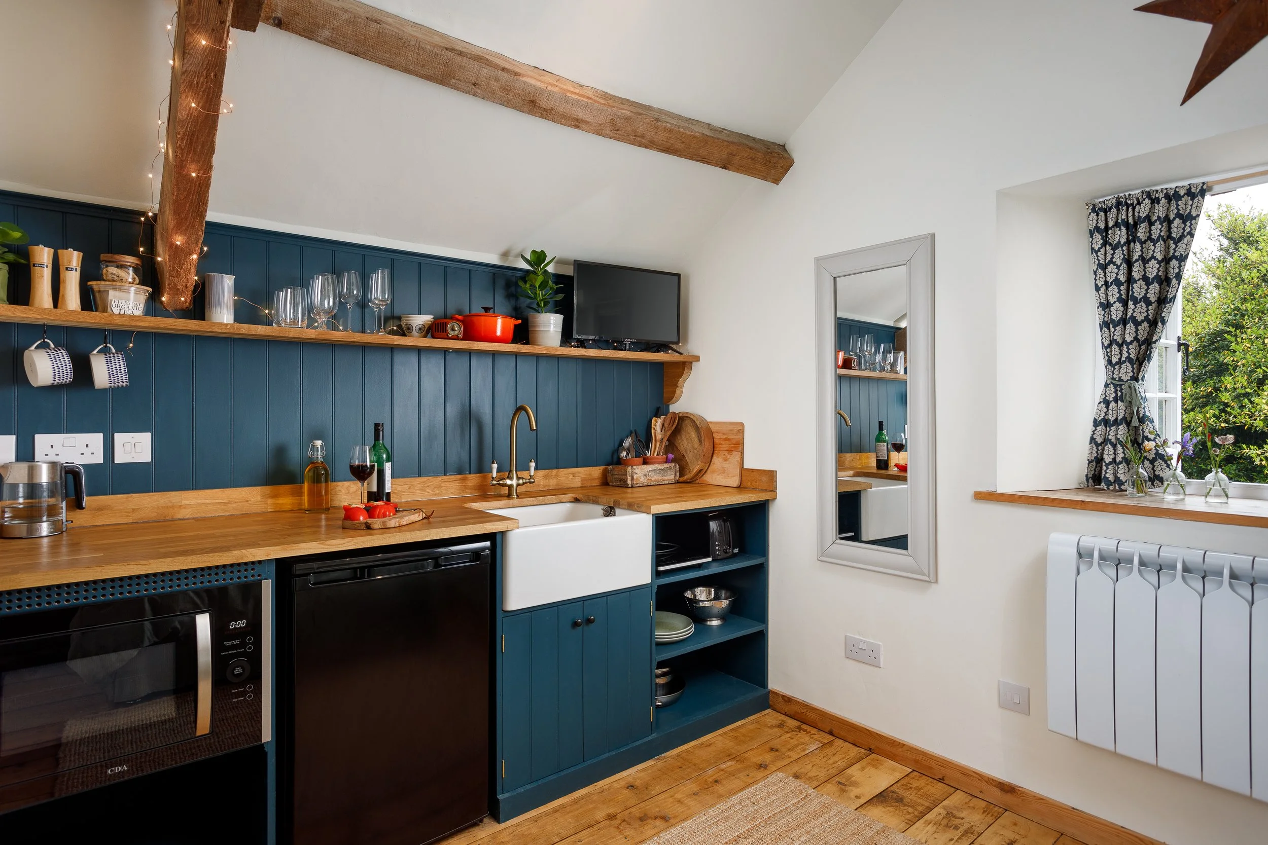 A cozy kitchen with dark blue lower cabinets, a wooden countertop, a white farmhouse sink, open shelves with glassware and dishes, a window with blue patterned curtains, and a mirror reflecting the kitchen interior.