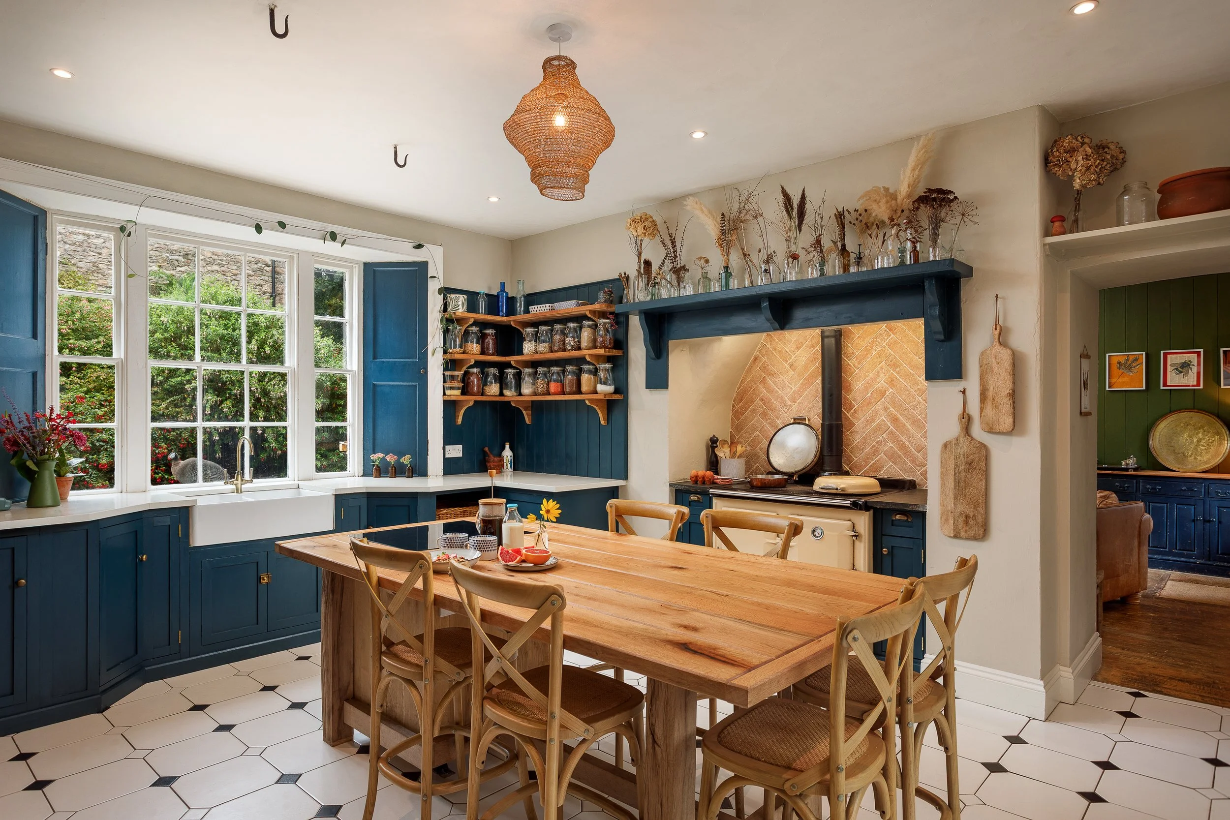 A cozy kitchen with blue cabinets, a wooden dining table with six chairs, a large window with open blue shutters, open shelves with jars and bottles, a fireplace with a brick arch and black pipe, and dried flowers and plants on the shelf and mantel.