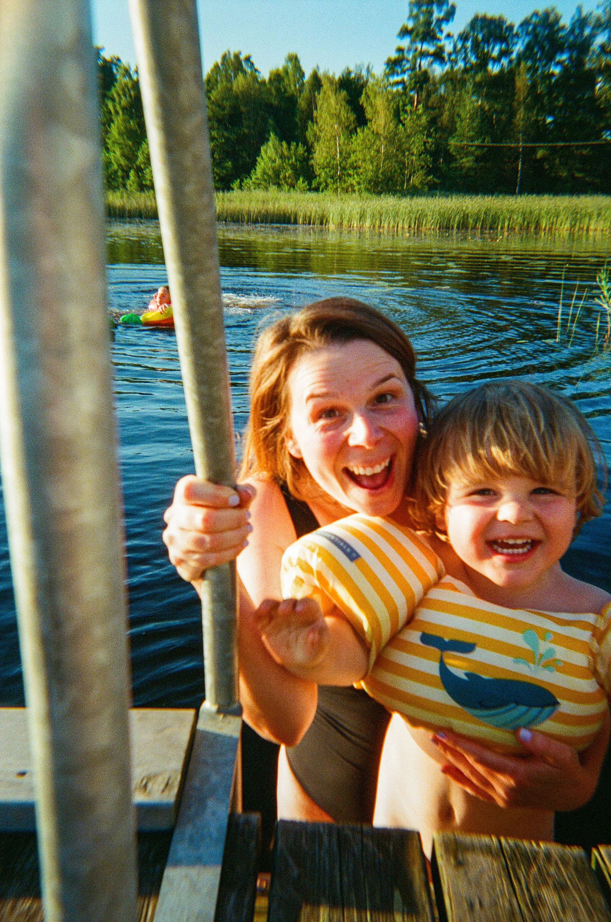 A woman and a young boy smiling and holding onto a ladder on a dock by a lake, with a swimmer in a tube and green trees in the background.