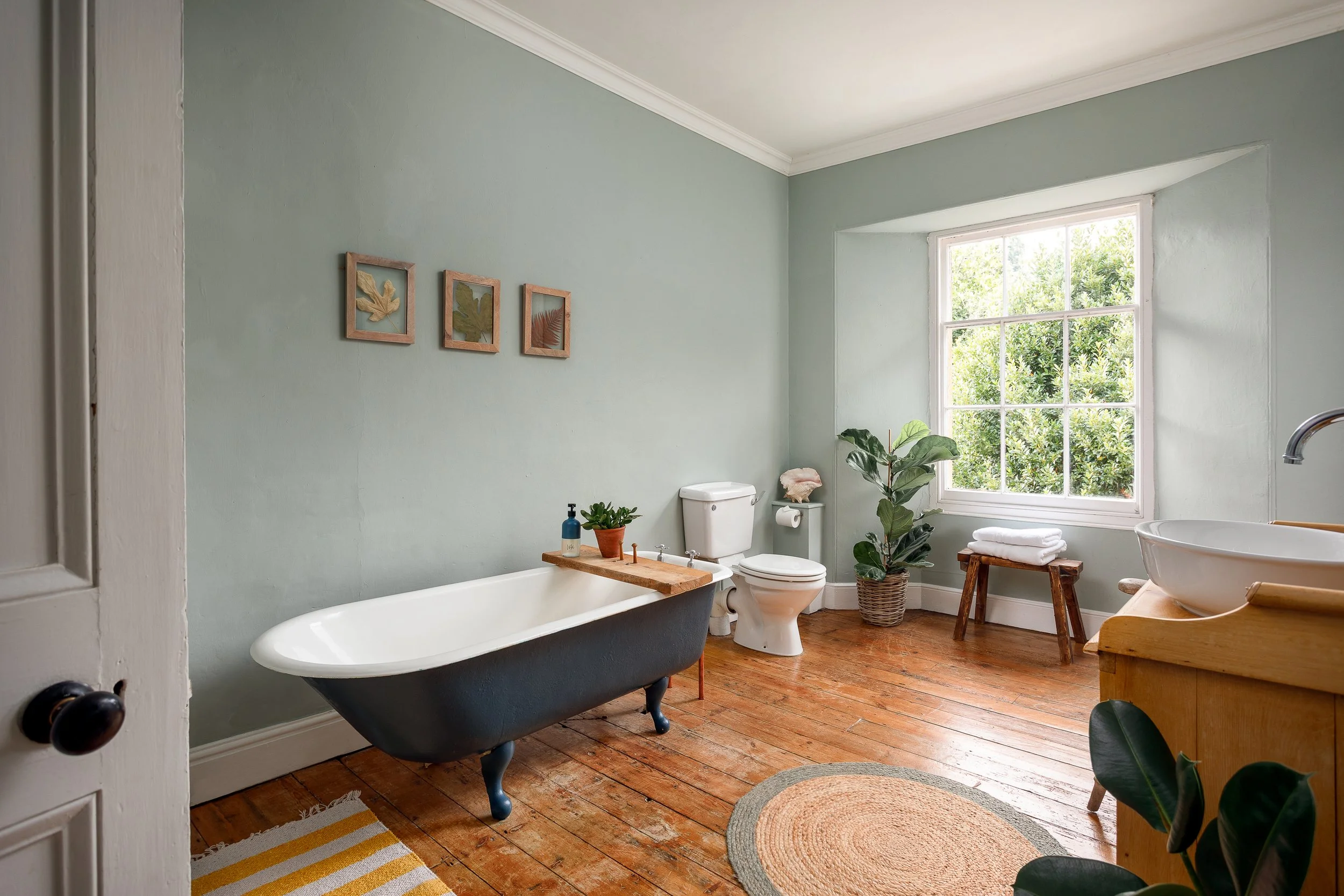 Bathroom with a clawfoot bathtub, toilet, window with greenery outside, wooden furniture, potted plants, and framed art on light green walls.