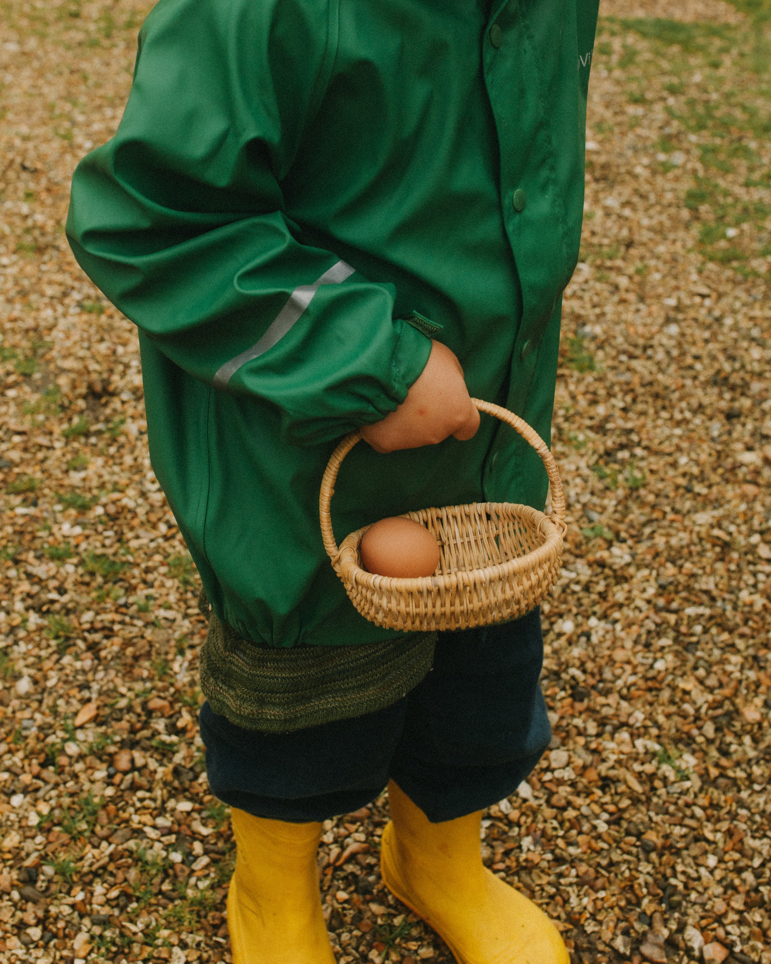 A child wearing yellow rain boots, a black skirt, a knitted sweater, and a green jacket holding a small wicker basket with an egg inside, standing on a gravel surface outside.