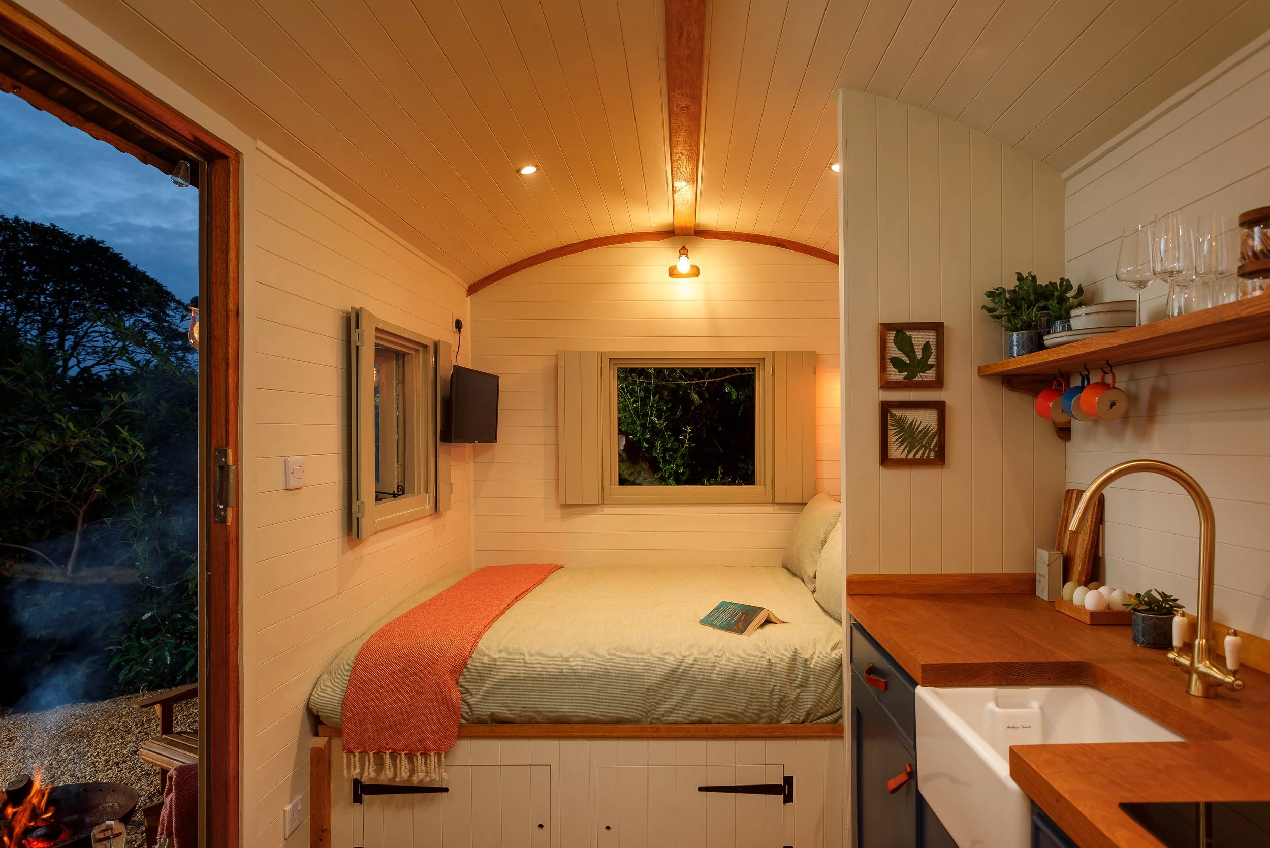 Interior of a cozy tiny home with a bed, small kitchen area, and a window showing night outside, warm lighting, wood panel walls and ceiling.