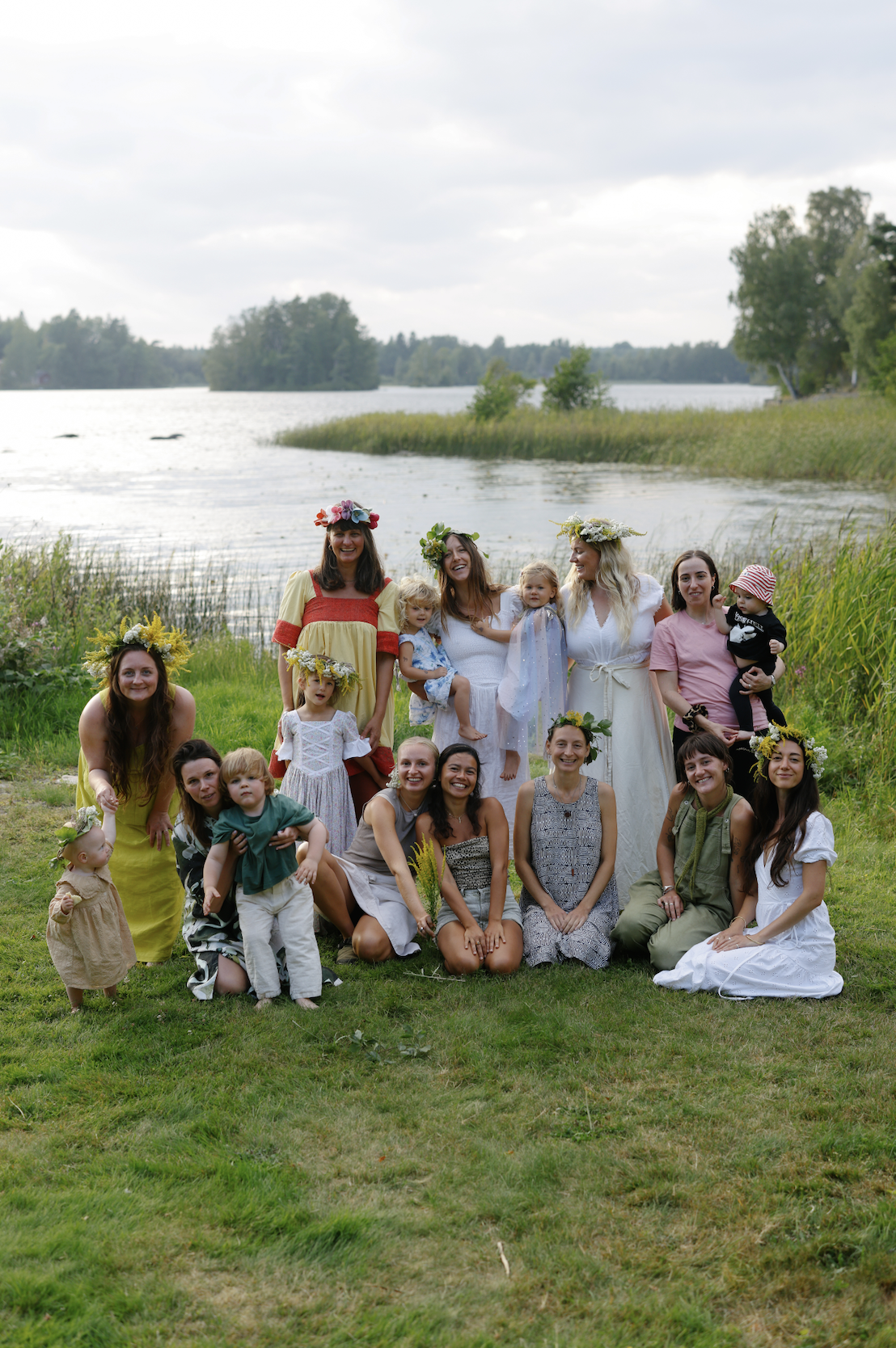 Group of women and children celebrating outdoors near a lake, some wearing floral crowns and dresses, in a natural setting with trees and water.