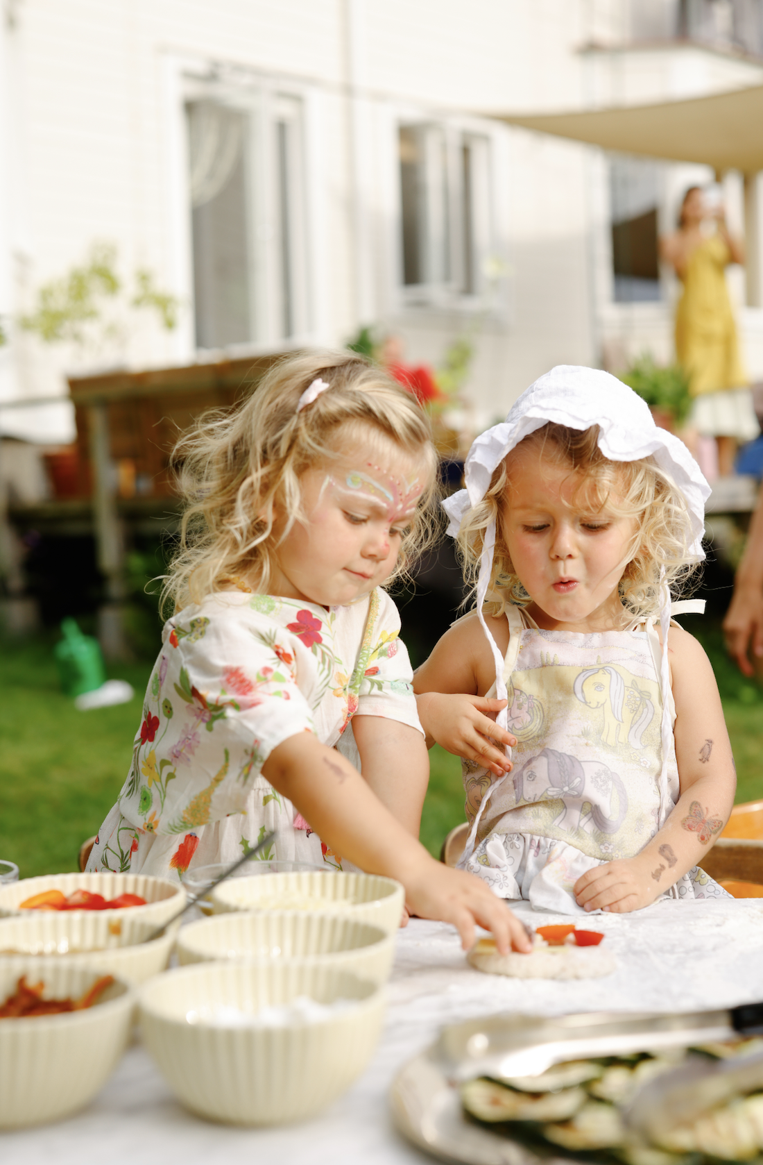 Two young girls with face paint and tattoos are making food together outdoors during a gathering, with various bowls and plates on the table.