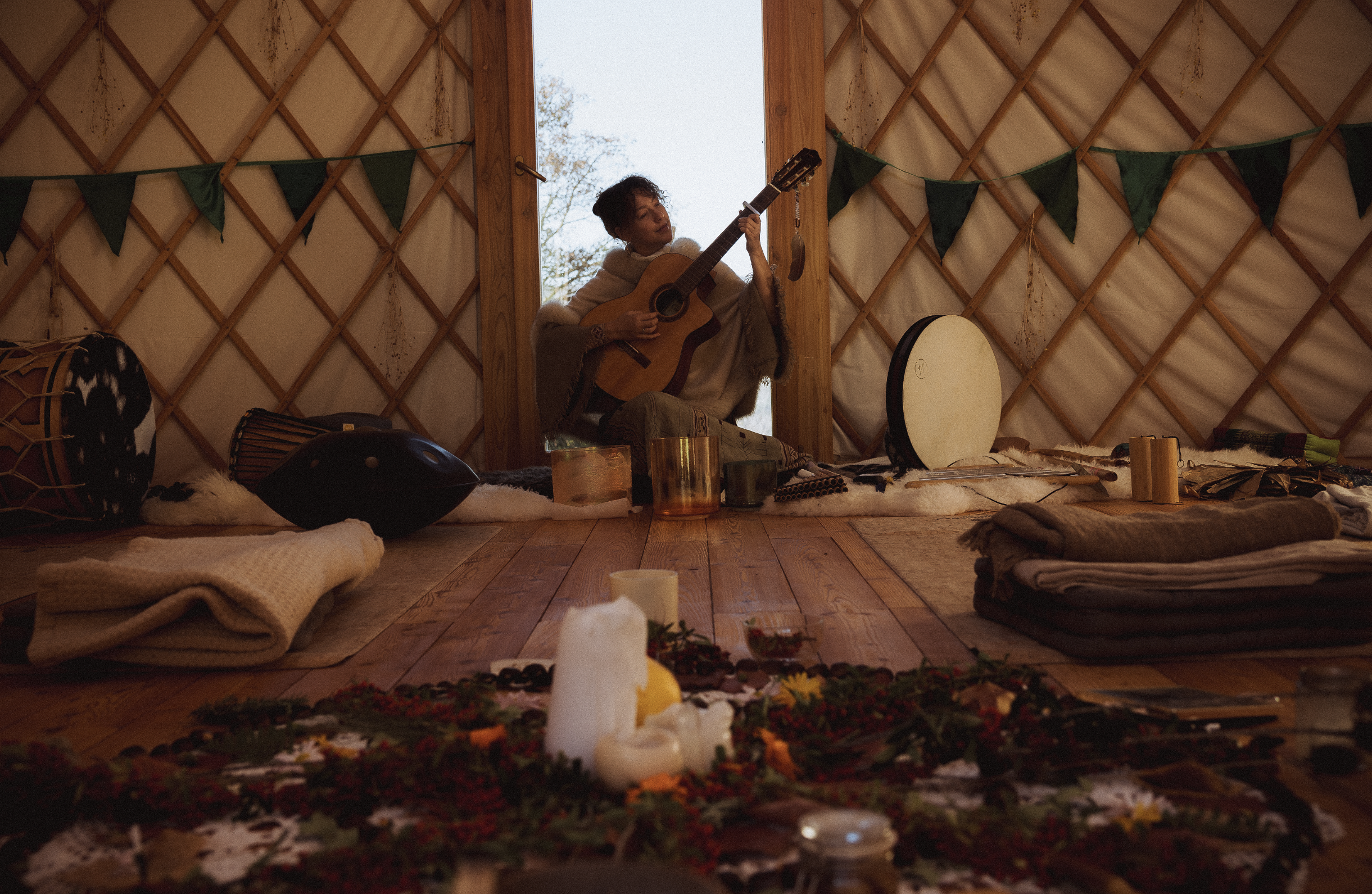 Person sitting in a yurt playing guitar, surrounded by musical instruments, candles, and cozy textiles, with natural light coming from the open door.
