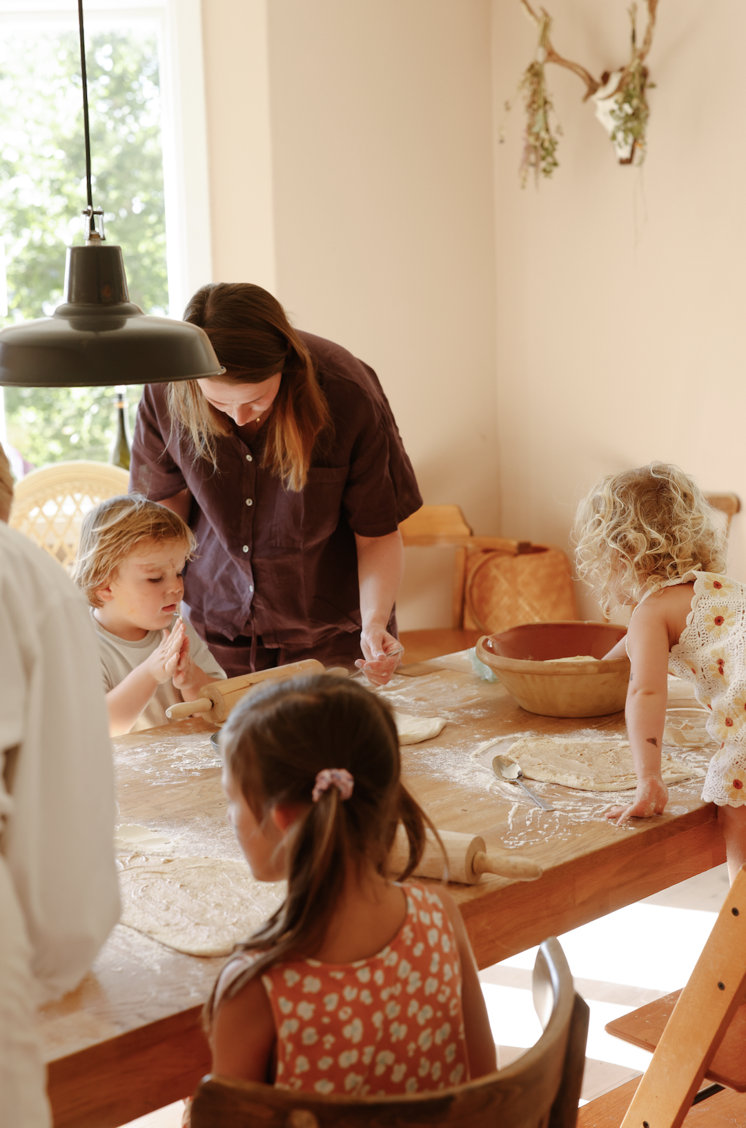 A woman and four children preparing dough on a wooden table, with children engaged in baking activities, in a bright kitchen with a window and hanging decorations.