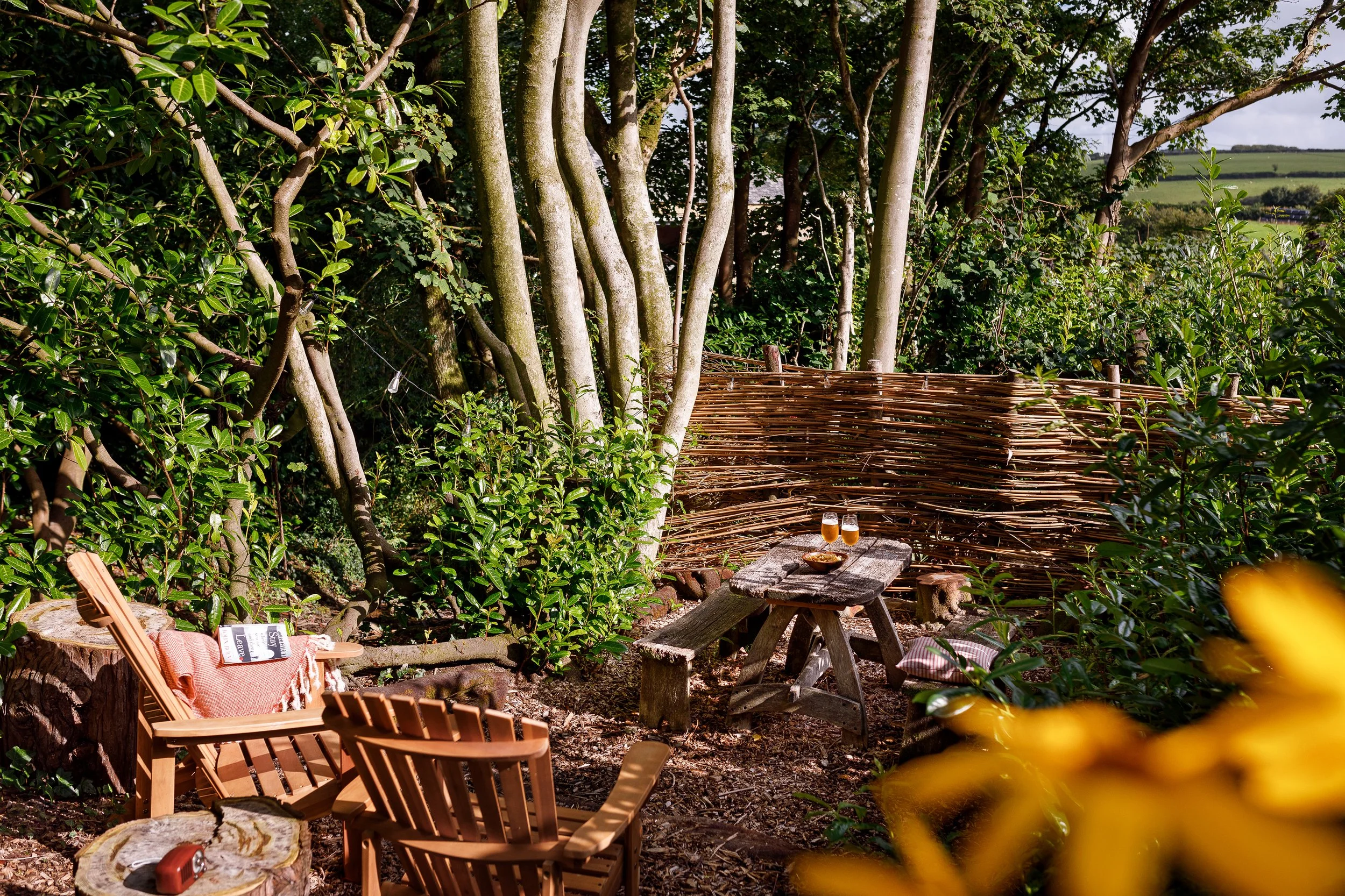 A cozy outdoor seating area in a garden with two wooden chairs, a small wooden table, and a woven fence backdrop, surrounded by lush green trees and bushes, with two glasses of beer on the table.