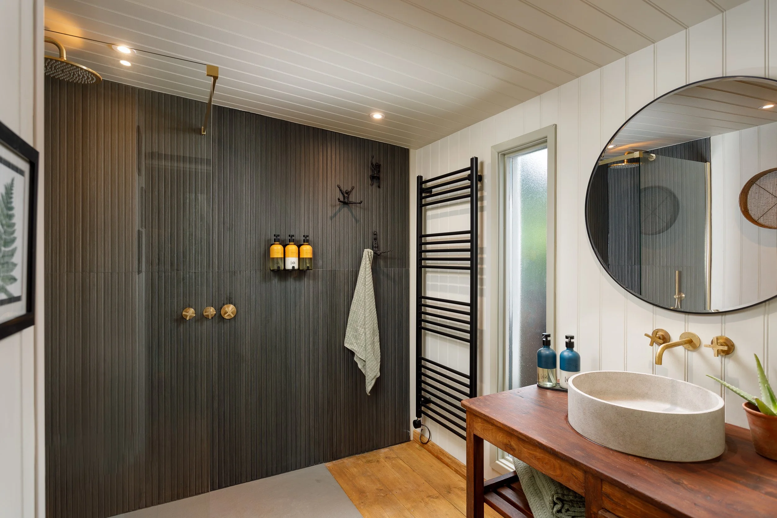 Modern bathroom with black textured wall, large round mirror, black towel rack, wooden vanity with stone sink, and black and yellow soap dispensers.