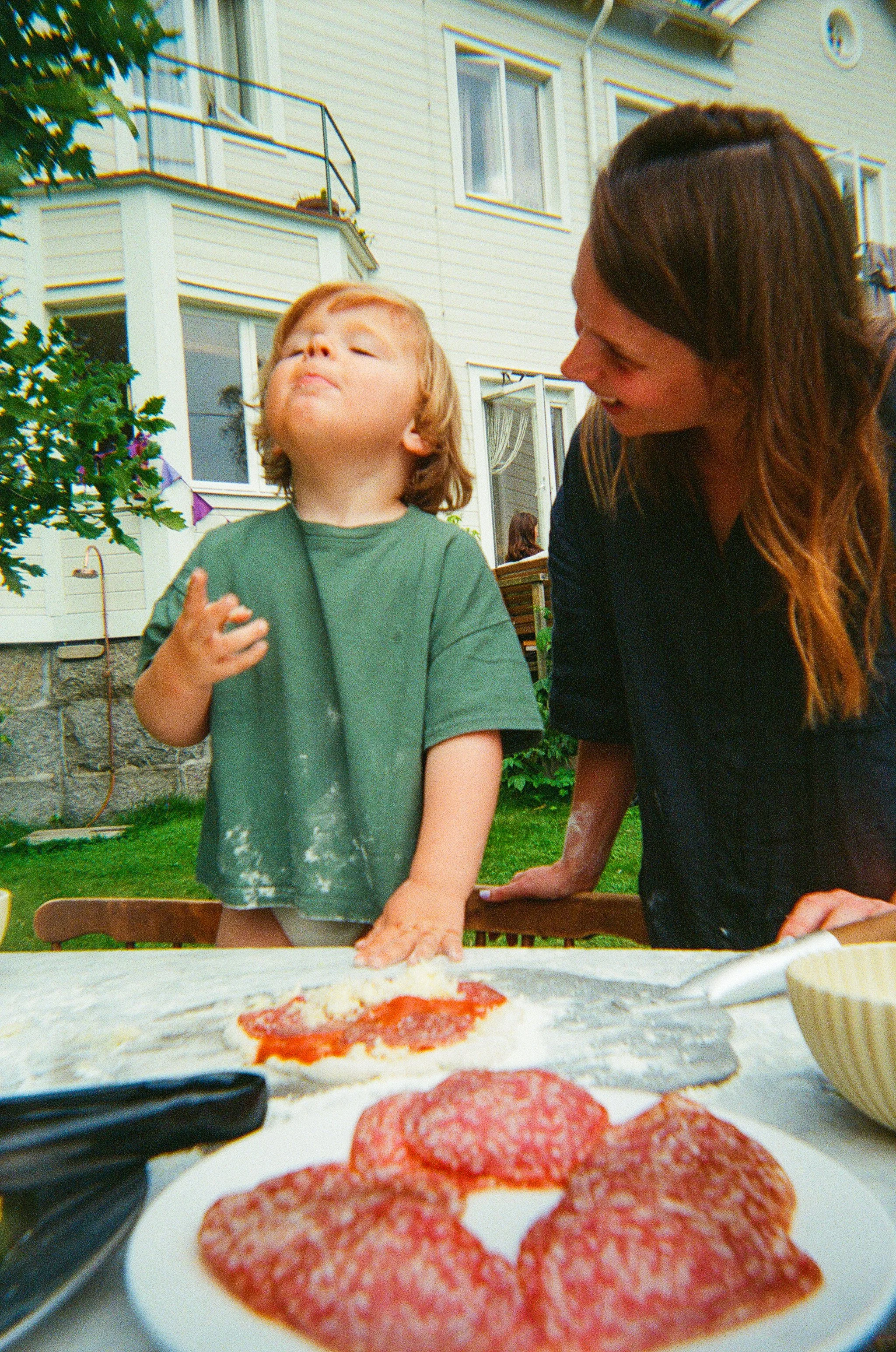 Child with flour on shirt and a woman smiling at him, making pizza outdoors with pizza ingredients on the table.