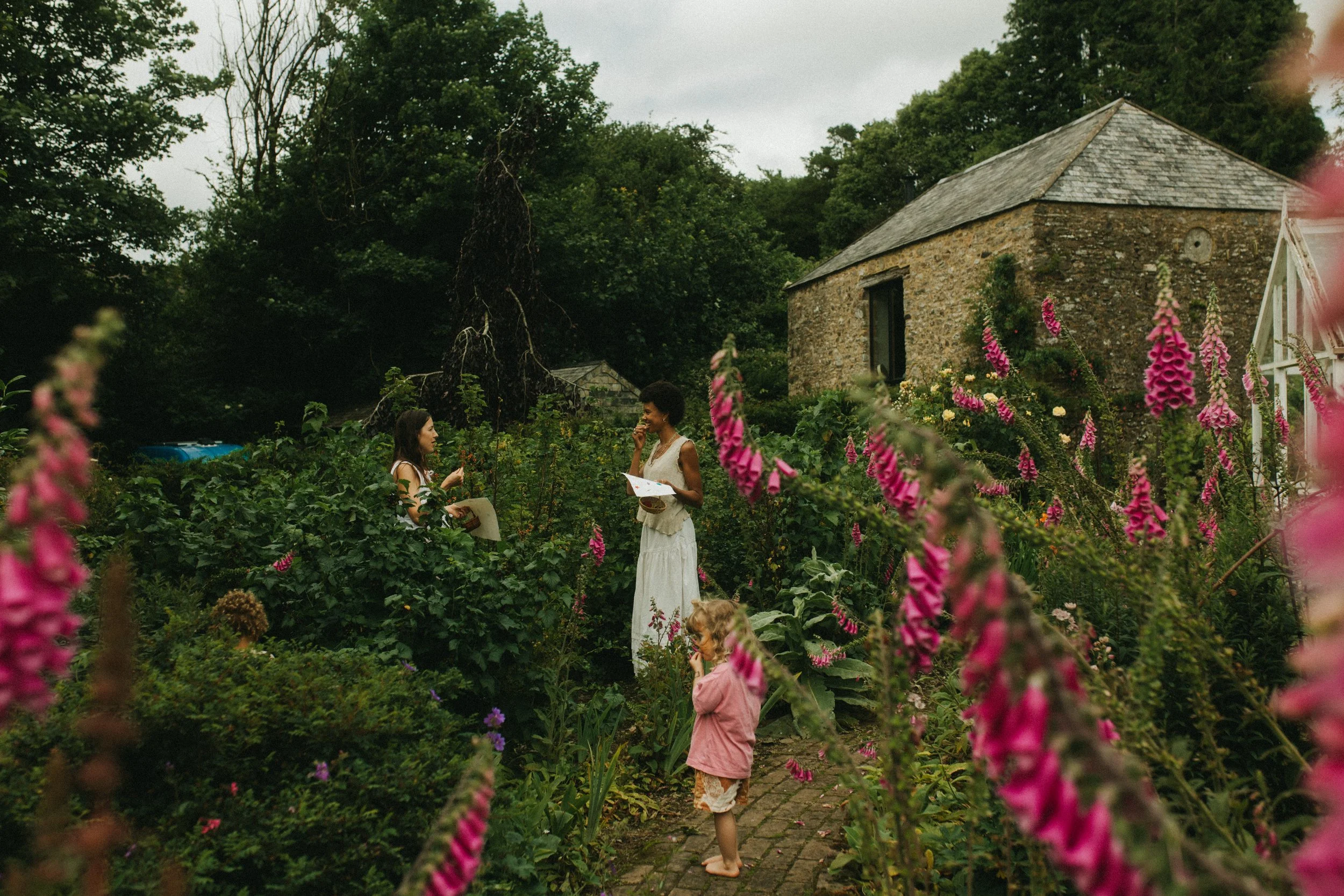 Two women and a young girl in a lush garden with tall pink flowers. The women are engaging in conversation, one with a notepad. There is an old stone building and trees in the background.