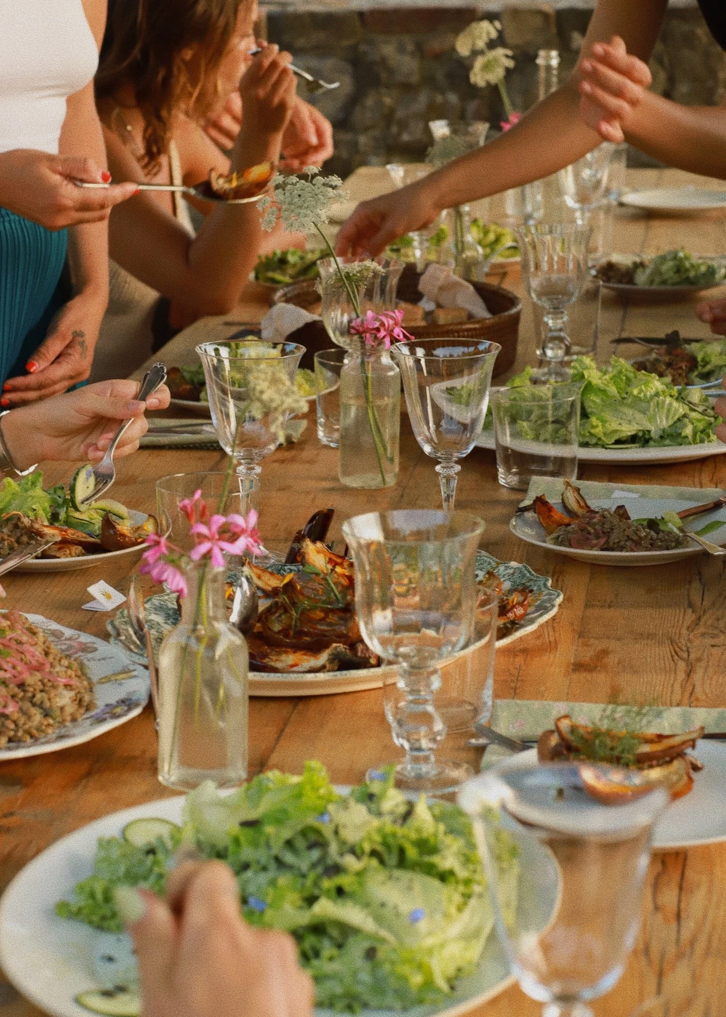 People having a meal at a long wooden table with dishes of salad, grilled meat, and drinks. Some individuals are serving themselves from bowls and plates, with floral decor and glasses on the table.