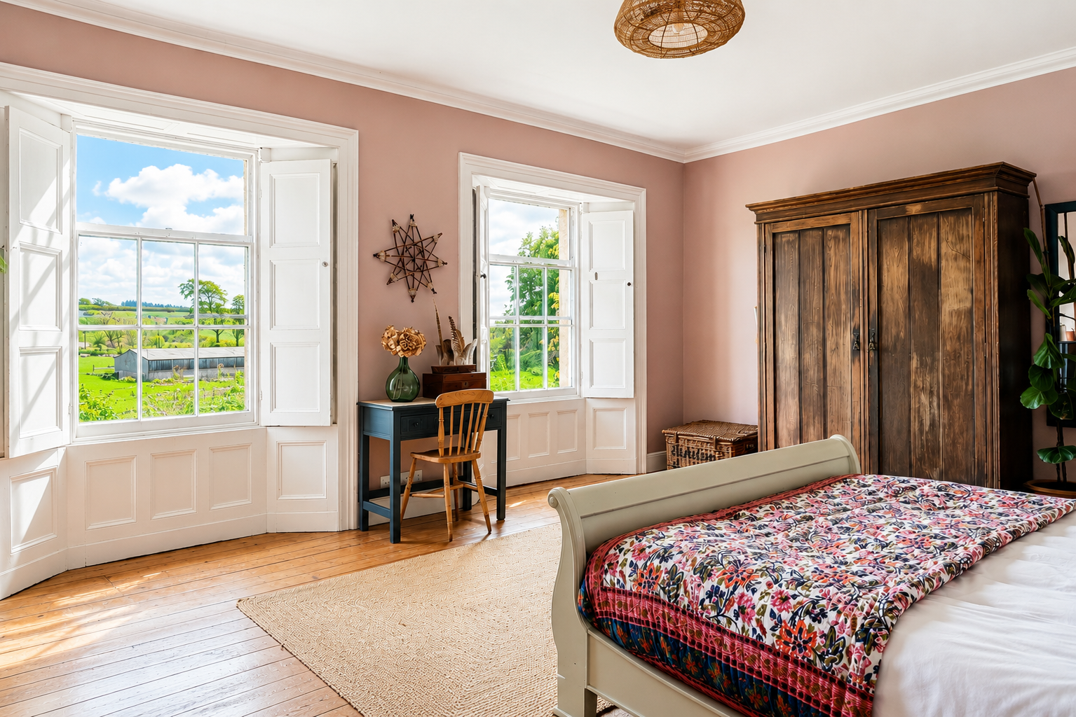 Bright bedroom with two open windows showing a farm scene with green fields and blue sky. There is a white bed with a colorful floral quilt, a dark wooden wardrobe, a small desk with a vase and flowers, and a wooden chair. The room has light pink walls and hardwood floors.
