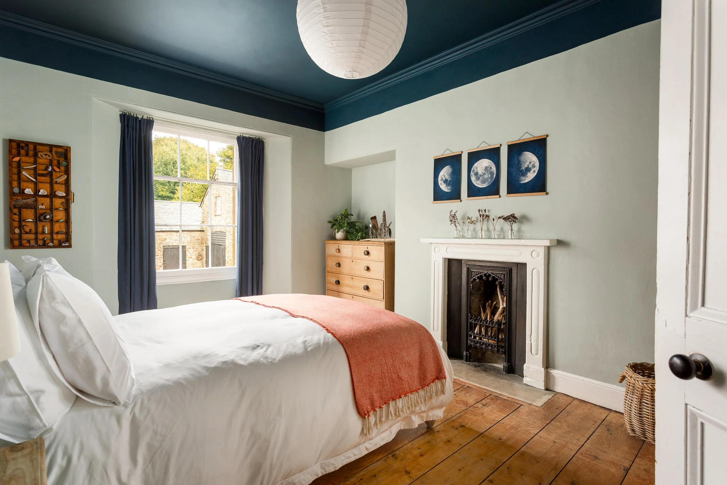 A cozy bedroom with a large bed, white linens, and an orange blanket at the foot. There is a wooden dresser with plants and decorative items on top, and a fireplace on the wall. The room has a large window with blue curtains, and three moon phase pictures above the fireplace. The ceiling is painted dark blue with a paper lantern hanging from it. The room has wooden flooring.