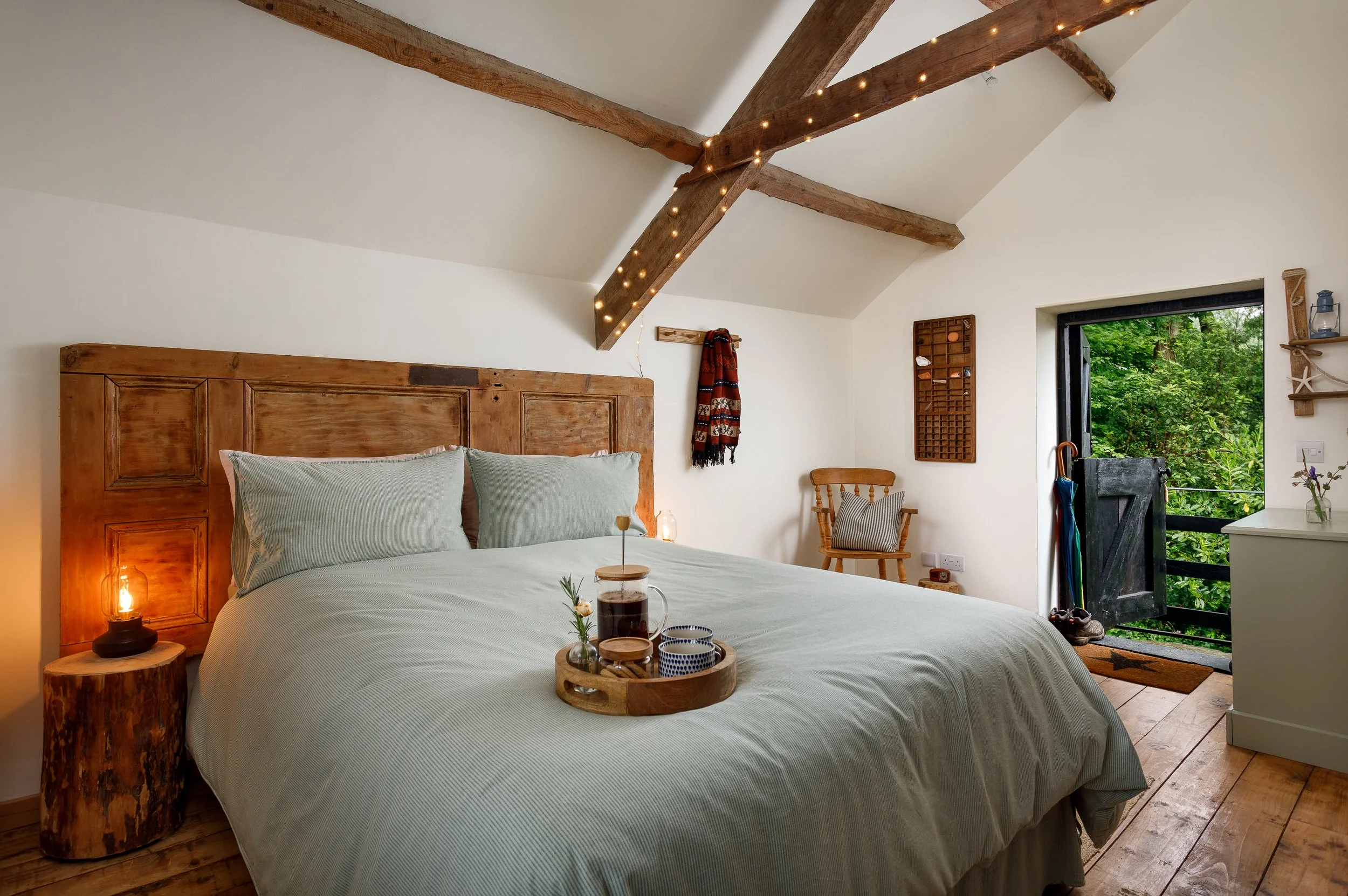 Cozy bedroom with a wooden bed frame and green bedding. A bedside table with a lamp, and a tray with drinks and flowers. Exposed wooden beams with string lights, a window showing greenery outside, and wall decorations.